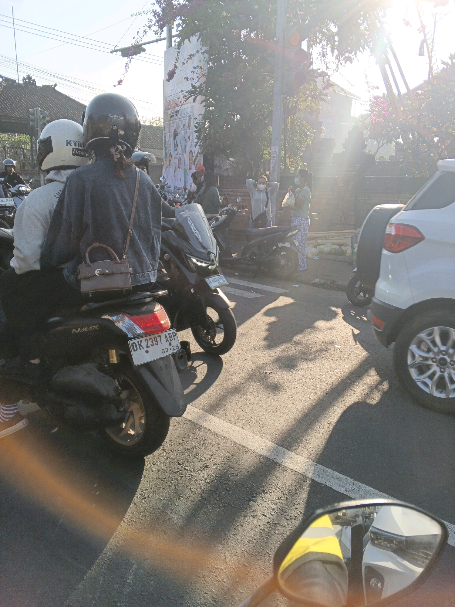 The image depicts a street scene in Abiansemal, Indonesia, during what appears to be a sunny day, possibly late afternoon due to the long shadows cast. Two people wearing helmets are on a black NMAX motorcycle, viewed from behind. The person in the front wears a white helmet and a grey jacket. The person in the back wears a dark helmet and a dark denim jacket. They are stopped in traffic, as indicated by the presence of other vehicles, including a white car on the right and several motorcycles. The license plate of the NMAX motorcycle reads "OK 2397 ABP".
In the background, several other people are visible. Two individuals are standing by the roadside, one wearing a light grey jacket and a face mask, the other wearing a green shirt and patterned pants, also with a face mask. They appear to be conversing or waiting. A large billboard or poster is mounted on a pole, featuring images of people. The environment suggests a developed urban or semi-urban area with overhead wires and traffic signals. The overall lighting condition indicates bright sunlight, creating lens flares and distinct shadows on the asphalt road. The image is taken from the perspective of another motorcycle.