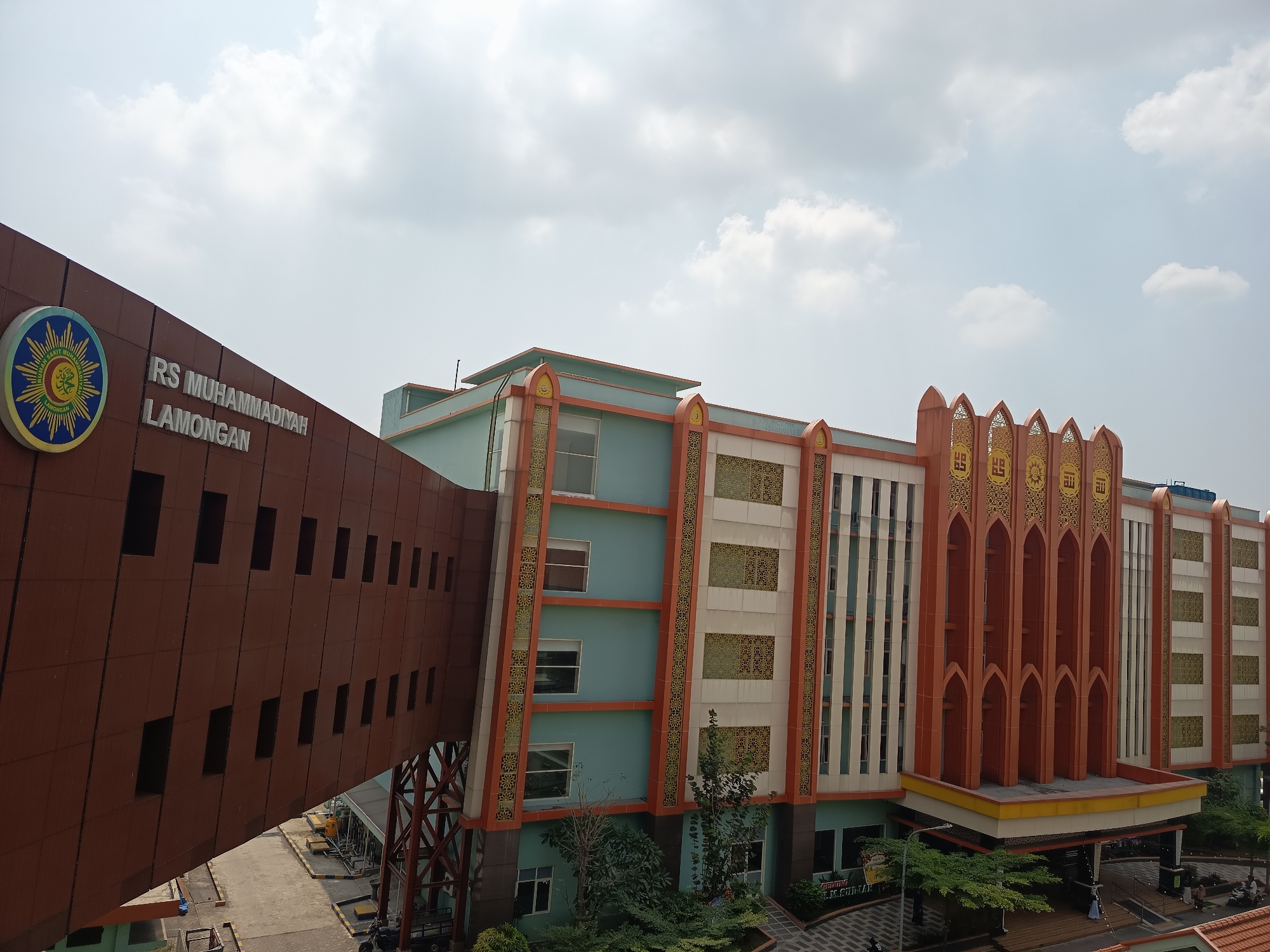 An exterior, eye-level, wide shot captures a modern hospital building with a skybridge in Dapur, Indonesia. The building is a multi-story structure featuring a light teal facade accented with orange trim and intricate gold patterned panels. A prominent, rust-colored skybridge extends from the left side of the frame, featuring a series of dark, rectangular windows and a large circular emblem with Arabic script and a sun-like design on its exterior. The skybridge is supported by a dark metal frame. The main building has a decorative, arched entrance with prominent vertical orange elements adorned with gold Arabic calligraphy. Lush green trees and landscaping are visible at the base of the building. The sky above is partly cloudy with a pale blue hue. A small section of paved ground with yellow and black striped markings is visible beneath the skybridge, with a few parked vehicles and people in the distance near the building's entrance.