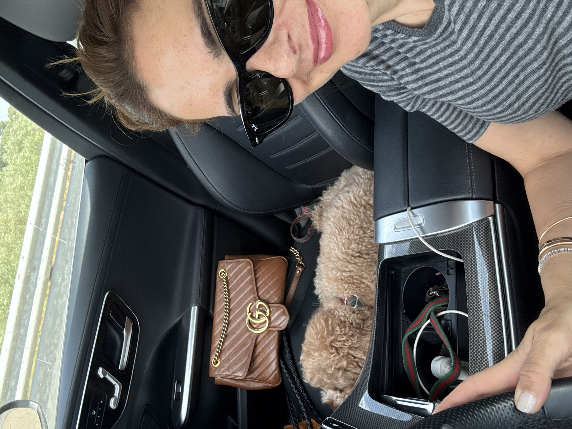An overhead self-portrait captures a woman and her small dog inside a car in Santarém, Portugal, during bright daylight. The woman, positioned in what appears to be the front passenger seat, wears black "Ray-Ban" sunglasses and a gray and white striped t-shirt. She has light brown hair, and her lips are painted with a glossy pink shade, suggesting a slight smile. Her right hand, adorned with stacked bracelets including one with diamonds, rests on the car's gear shifter or center console, revealing light-colored nail polish.
Beside her, a small, fluffy, light brown dog, possibly a poodle or similar breed, is nestled between the car seats. A brown quilted handbag with a prominent gold "GG" logo (Gucci) and a gold chain strap rests on the seat next to the dog. The car interior features black leather seats with intricate stitching, and the center console is accented with silver and carbon fiber trim. A compartment in the console is visible, containing charging cables, a small white object, and a red and green striped ribbon, reminiscent of Gucci branding.
Through the car window, green trees and a road with guardrails can be seen, indicating the vehicle is either driving or parked beside a route. The overall scene suggests a relaxed moment during a car journey.