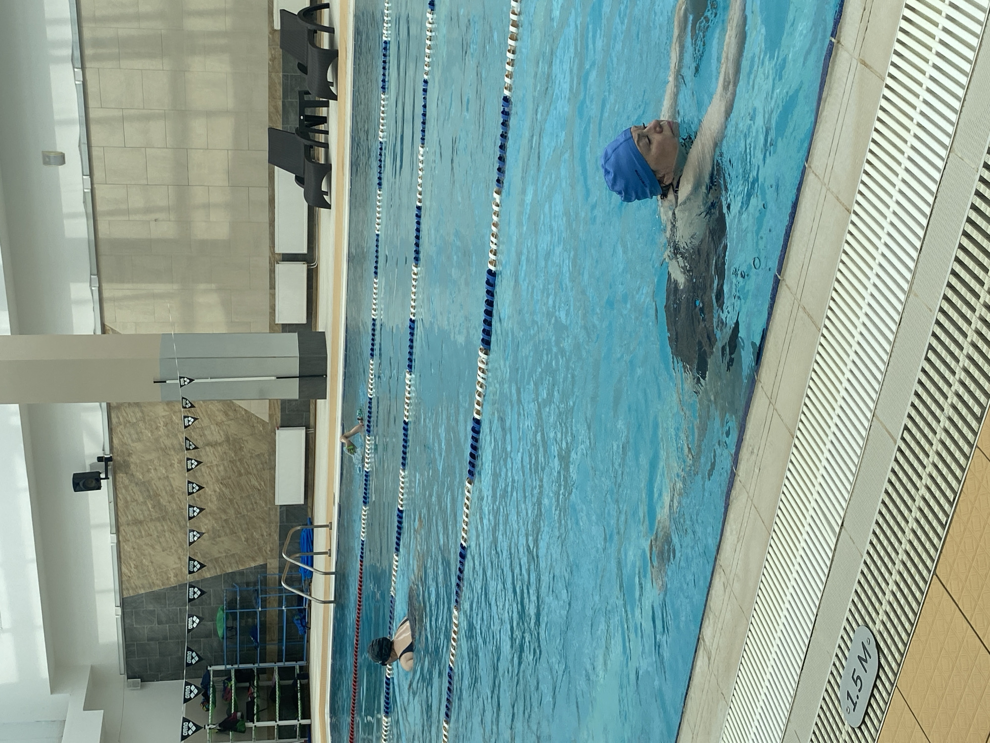The image shows an indoor swimming pool. In the foreground, a woman wearing a blue swimming cap and dark swimsuit is swimming breaststroke. Further back, another person in a dark swimsuit and black swimming cap is also swimming. In the background, there are lane dividers and lounge chairs visible by the side of the pool. The architecture of the pool area suggests a modern facility, with tiled walls and ceiling lighting. There is a sign indicating "1.5 M" near the edge of the pool. There is no information in the image to suggest the location is Yekaterinburg, Russia.