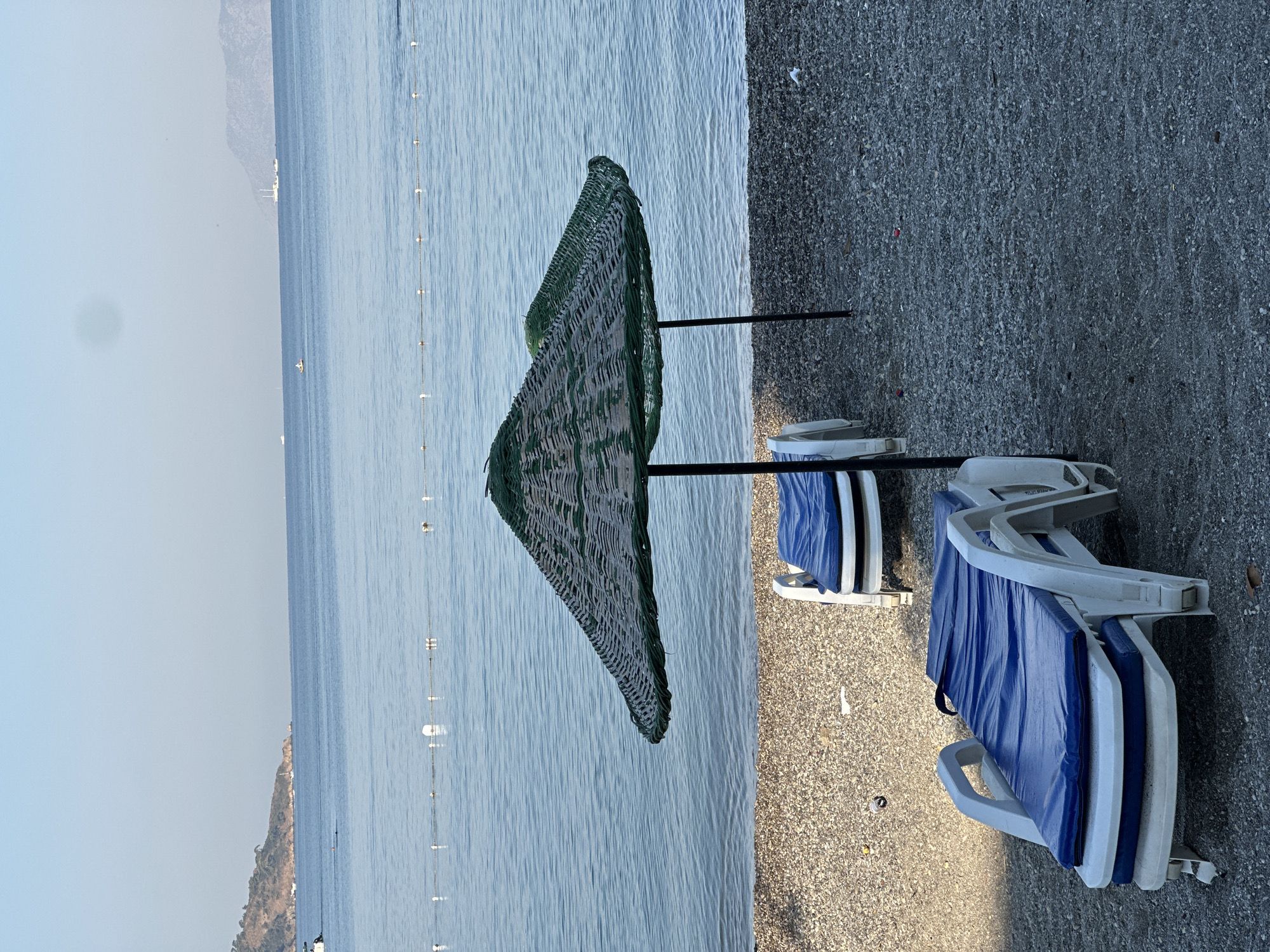 A beach scene in Fethiye, Turkey, features two empty lounge chairs with blue cushions on a pebble-strewn beach, positioned in front of two woven straw umbrellas with green trim. The umbrellas are supported by black poles. Beyond the beach, the calm, blue sea stretches to the horizon, with a few boats visible in the distance. A line of buoys marks a boundary in the water. The sky is clear and pale blue, with a distant, hazy coastline to the left and mountains in the far distance. The overall impression is one of quietude, with the beach appearing unoccupied.
