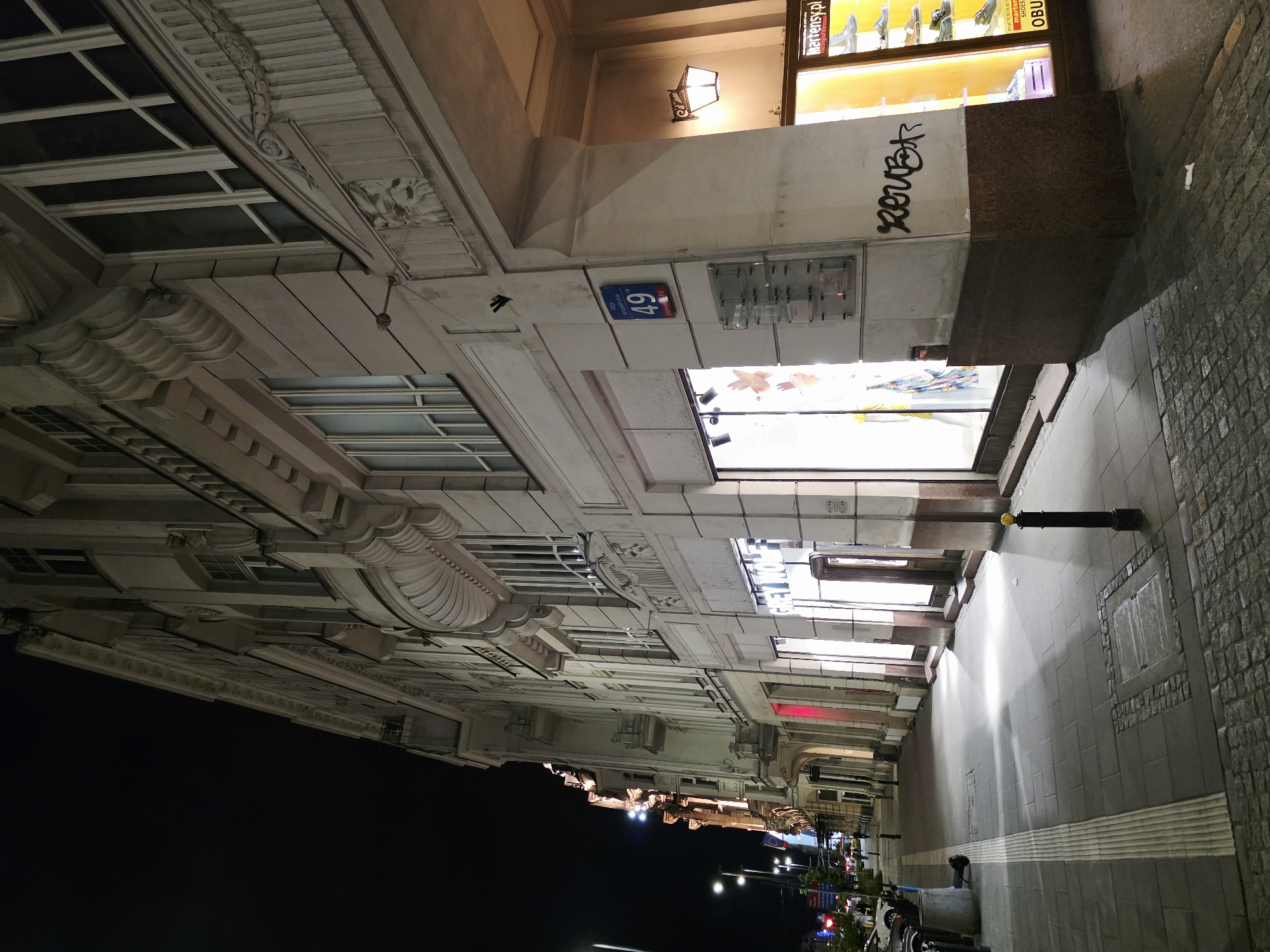 The image depicts a street scene at night in Warsaw, Poland, specifically on Marszałkowska Street. The perspective is from the sidewalk, looking down the street alongside a large, ornate building with shops on the ground floor. The building's facade is light-colored, possibly stone or stucco, and features classical architectural details like columns, cornices, and decorative carvings. Several windows are visible on the upper floors, some with ornate grilles.
The ground floor houses commercial establishments. One storefront, labeled "GREY WOLF," is brightly illuminated, showcasing mannequins or displays of clothing. Another shop, further to the right, has a brightly lit display window featuring shoes, with text that appears to say "Martensy.pl" and "The Original Dr. Martens." A street sign with the number "49" and Polish text indicating a street name is affixed to the building.
The sidewalk is paved with large grey tiles and features a tactile paving strip for the visually impaired. A black bollard with a yellow top stands on the sidewalk. Streetlights cast pools of light on the pavement. In the distance, the street continues, with more illuminated buildings and traffic visible. A few cars are parked along the street. There are no people clearly visible in the foreground, though figures can be seen further down the street. The overall atmosphere is that of a city at night, with urban infrastructure and commercial activity. Graffiti is visible on the lower part of the building near the shoe store.