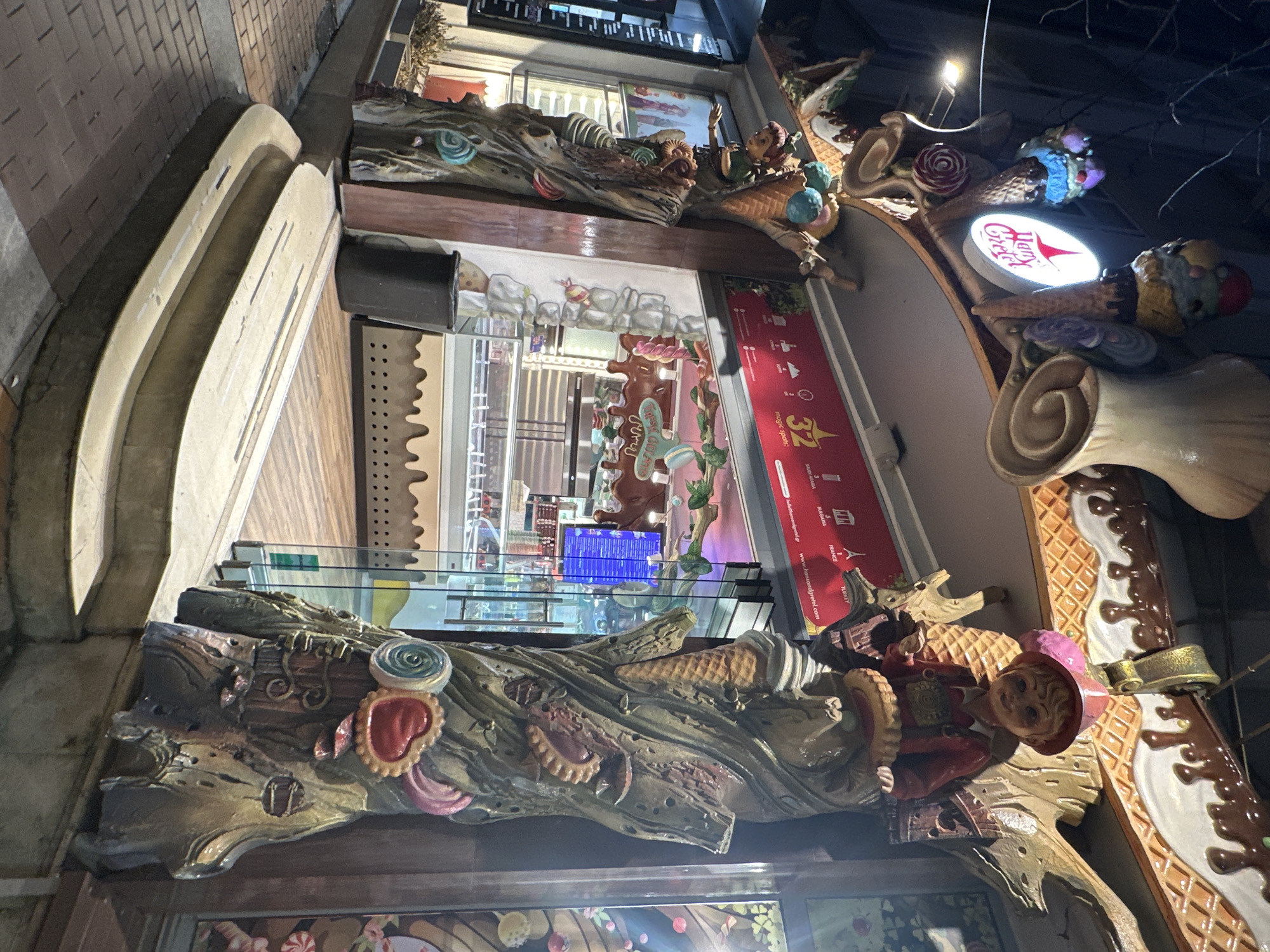The image depicts a facade of an ice cream shop. The architecture is elaborate and whimsical, with decorations of ice cream cones, cookies, and fairytale-esque figures crafted into the structure. The shop's name, "Vanila Kitchen," is illuminated by a sign. Display cases showcase a variety of ice cream flavors and desserts. A display screen in the back shows a blue display. The store is located in the city of Sofia, Bulgaria, as indicated by the presence of Cyrillic script on a sign.