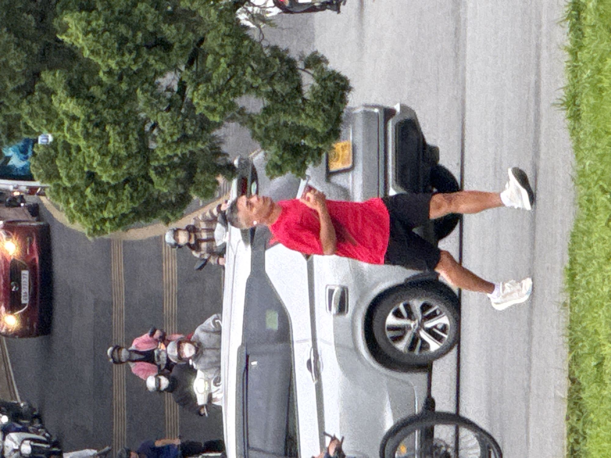 A man in a red t-shirt and black shorts is jogging on a paved road. He is wearing white athletic shoes and white socks. A silver SUV is parked to his left. A large green tree is in the background, obscuring parts of the scene. Several motorcycles and people are visible further down the road, with some wearing face masks. The scene appears to be in an urban environment.