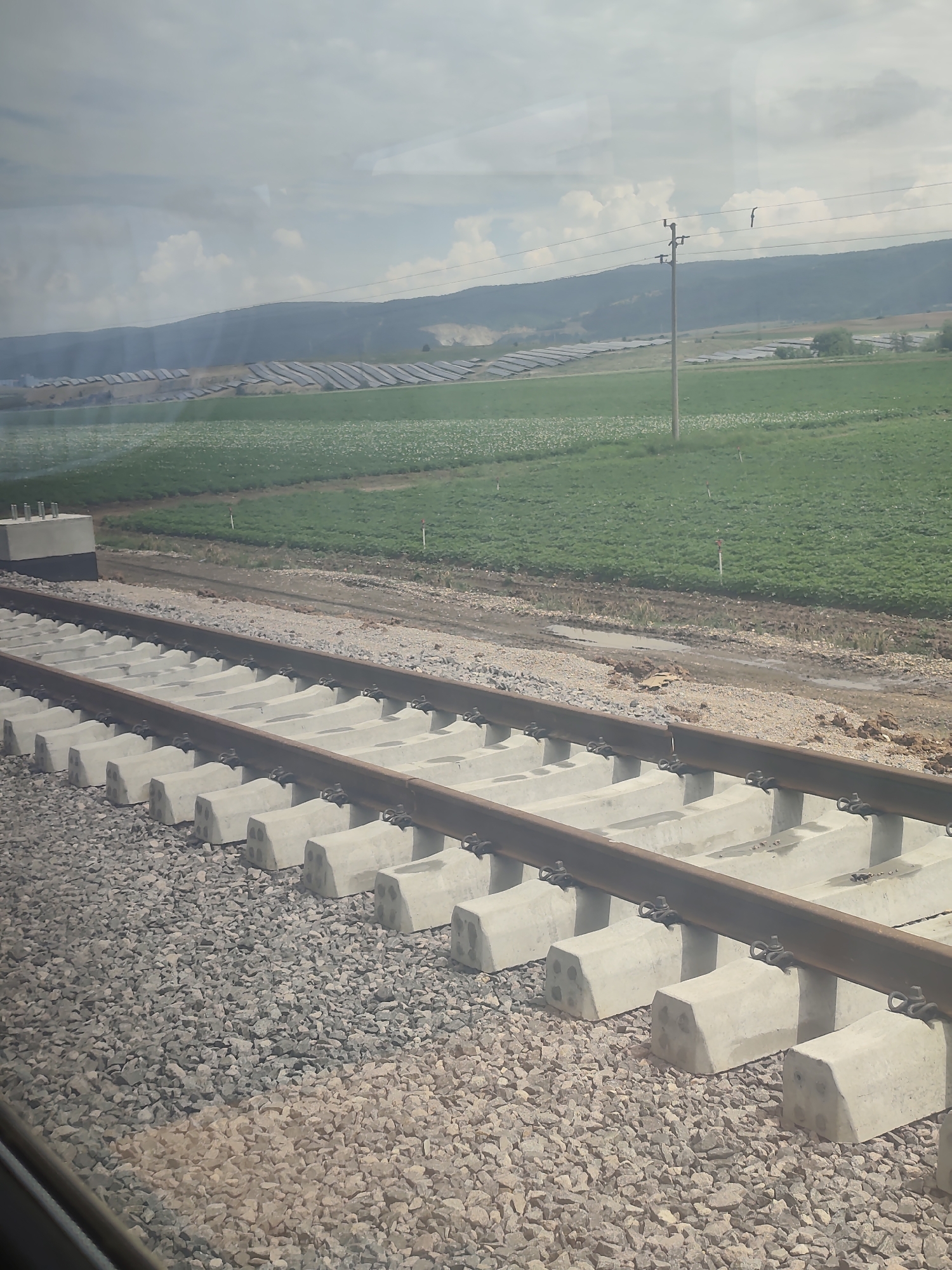 This image shows a new railway track under construction. The tracks are laid on concrete sleepers, which are embedded in a bed of gravel. In the background, there are fields of green crops, solar panels, and rolling hills under a cloudy sky. A single utility pole with wires is visible in the mid-ground, with two birds perched on the wires. The scene appears to be a rural or semi-rural area, possibly a construction site for infrastructure development. The lighting suggests it is daytime, with the sun possibly behind the clouds. There is no visible text in the image.