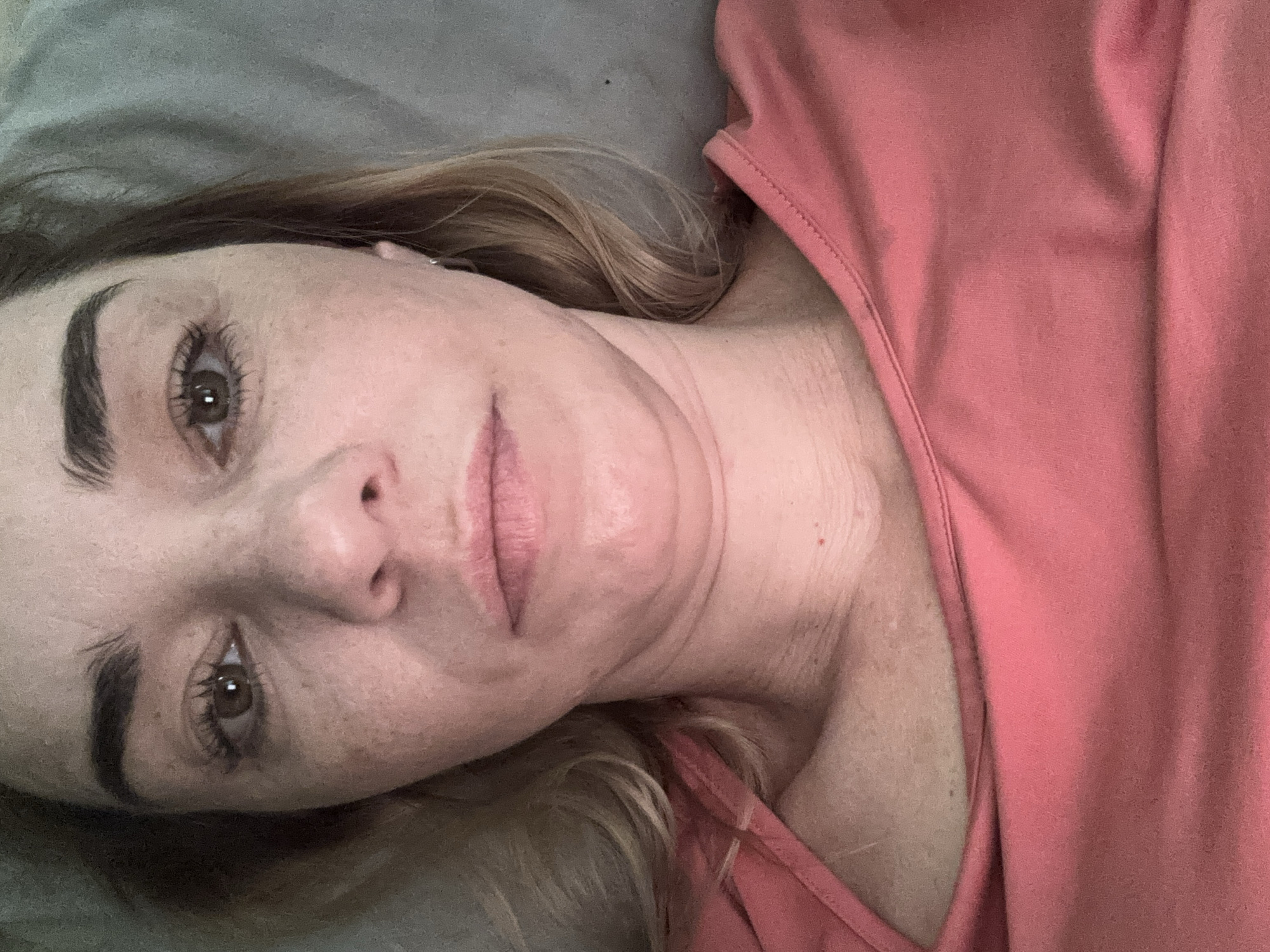 A person is lying down, facing the camera. Their head is tilted slightly. They have fair skin, brown eyes, and blonde hair. They are wearing a pink shirt. The background appears to be a gray surface, possibly a pillow or blanket. There is no information in the image that would indicate it was taken in Ladozhskoe Ozero, Russia, or any other specific location.