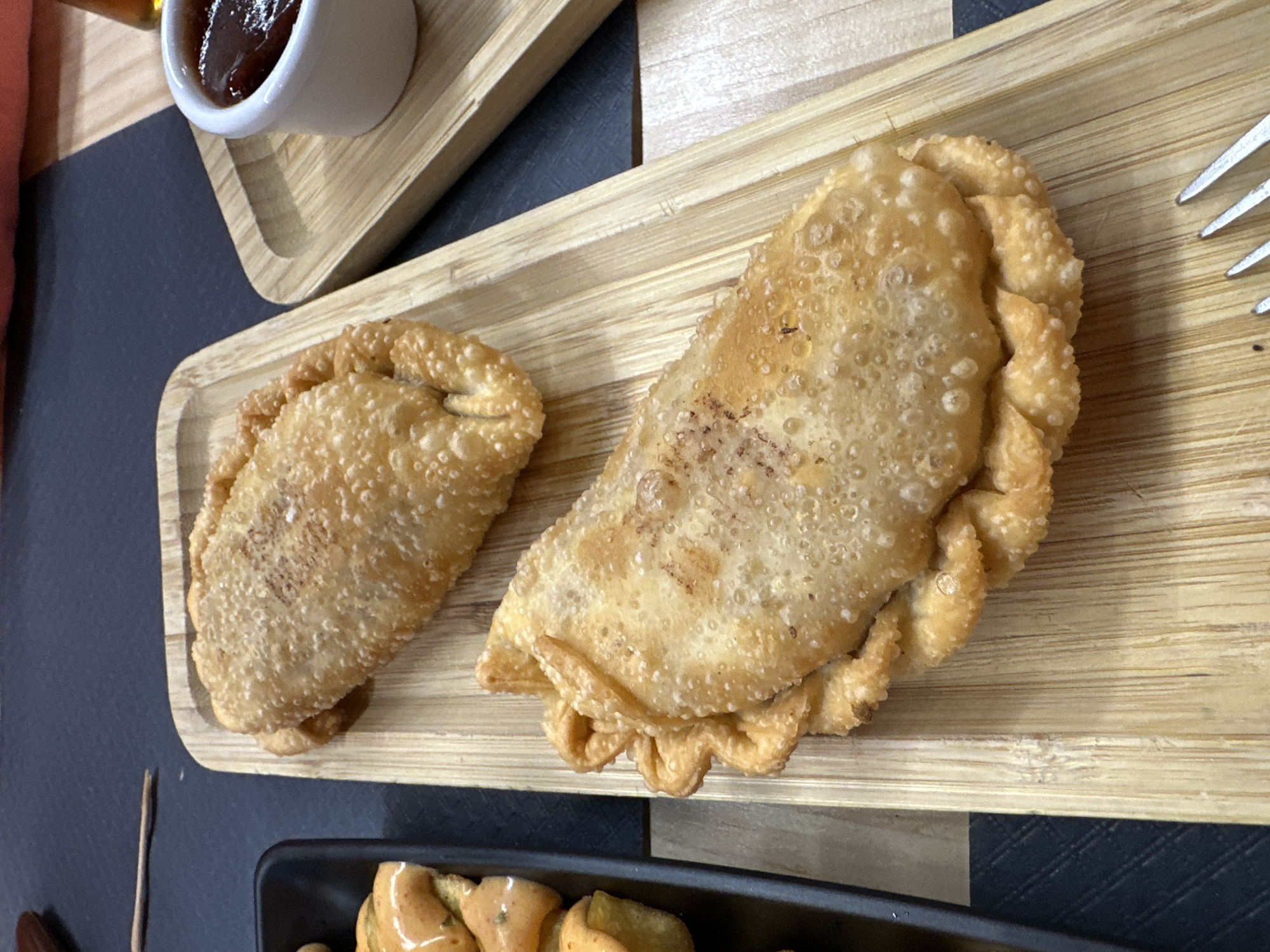 A close-up, high-angle shot showcases two golden-brown empanadas resting on a light wooden tray, likely in Encamp, Andorra. The larger empanada, on the right, appears to be freshly fried with a bubbly, textured crust. Its edges are crimped in a decorative fashion. A slightly smaller empanada sits to its left, displaying a similar fried, textured surface.
In the upper left corner, a small white ramekin filled with a dark, reddish-brown sauce or jam is visible on another section of the wooden tray. A silver fork with multiple tines is partially visible on the right side of the main wooden tray, suggesting a meal is either in progress or about to begin. The background consists of a dark, possibly black, tabletop with subtle texture. Another dark tray or dish, containing what appears to be more food or garnish, is partially visible at the bottom of the frame. The overall scene suggests a casual dining experience, focusing on traditional fried pastries. There is no visible text in the image.
