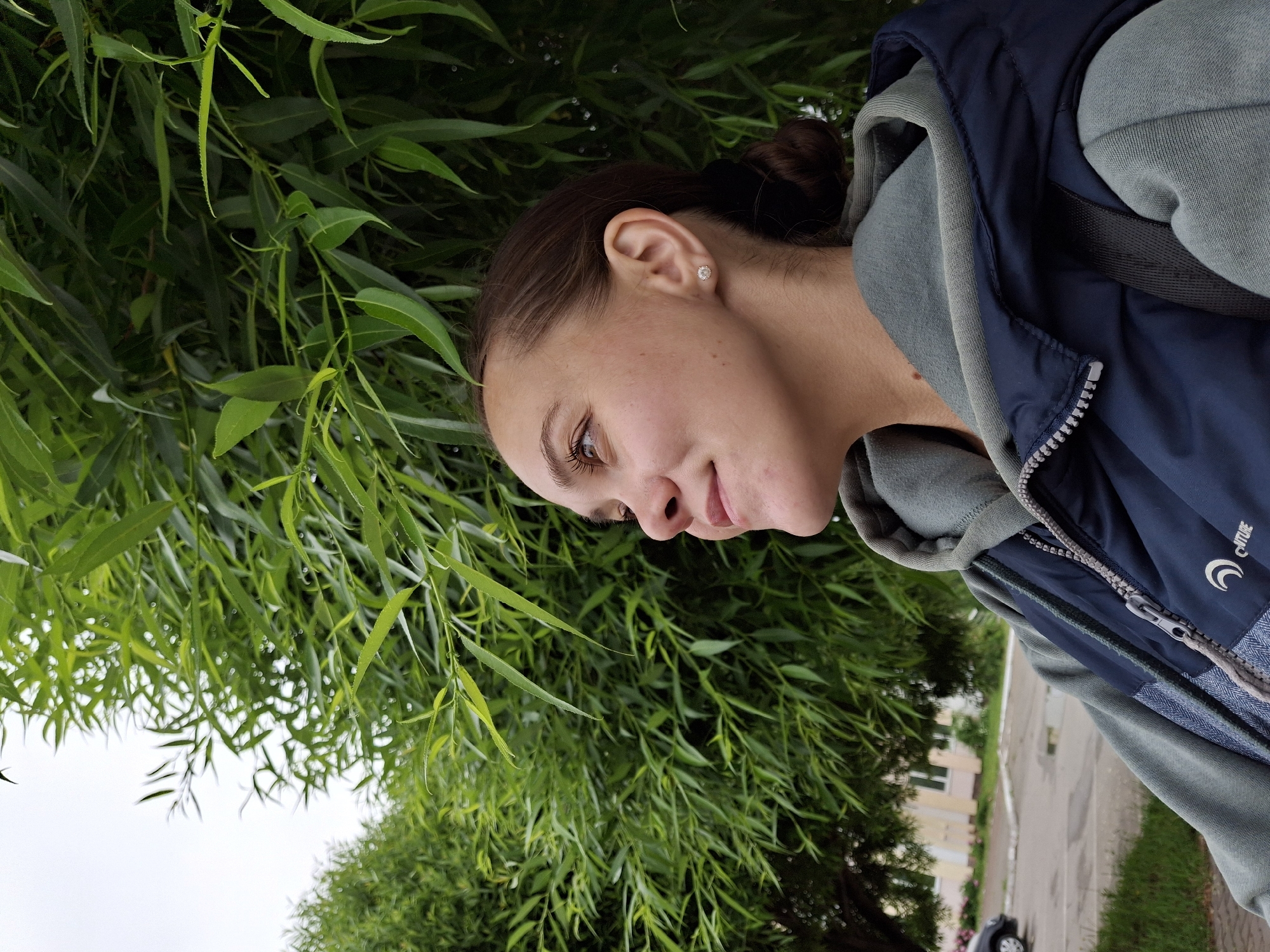 A young woman with light brown hair tied back in a bun is pictured from the chest up, looking to her left. She has a slight smile and is wearing a light grey hooded sweatshirt under a navy blue vest with a zipper. Her left ear is visible, adorned with a small stud earring. The background is dominated by lush green foliage, appearing to be willow branches, against an overcast sky. In the blurred background to the left, a street with a parked car and buildings can be discerned, suggesting an urban setting. This scene is consistent with outdoor conditions in Chaikovsky, Russia, particularly during warmer months when trees are in full bloom.