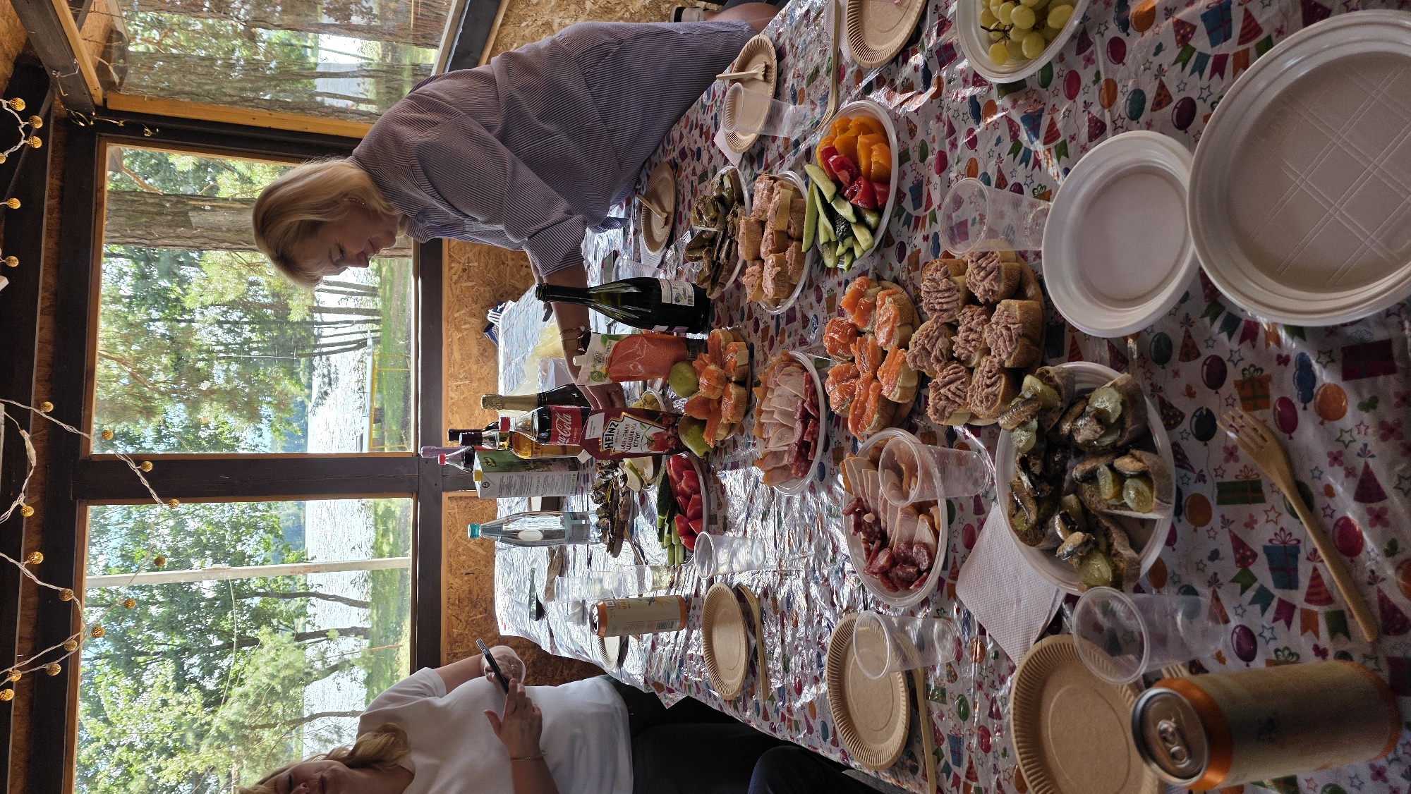 The image captures a gathering at an outdoor covered dining area, likely located in the Тэльмінскі сельскі Савет region of Belarus, overlooking a body of water surrounded by trees. A long table is laden with a variety of food and drinks, including an array of sandwiches, meats, sliced vegetables, a can of soda, and bottles of beverages. Two women are visible; one on the left in a white t-shirt holding a mobile phone, and another on the right wearing a striped shirt, actively placing food on the table. The atmosphere suggests a casual meal or picnic, with disposable plates and cups also present. The background reveals a natural landscape with a lake and dense foliage, implying a tranquil, possibly rural setting.