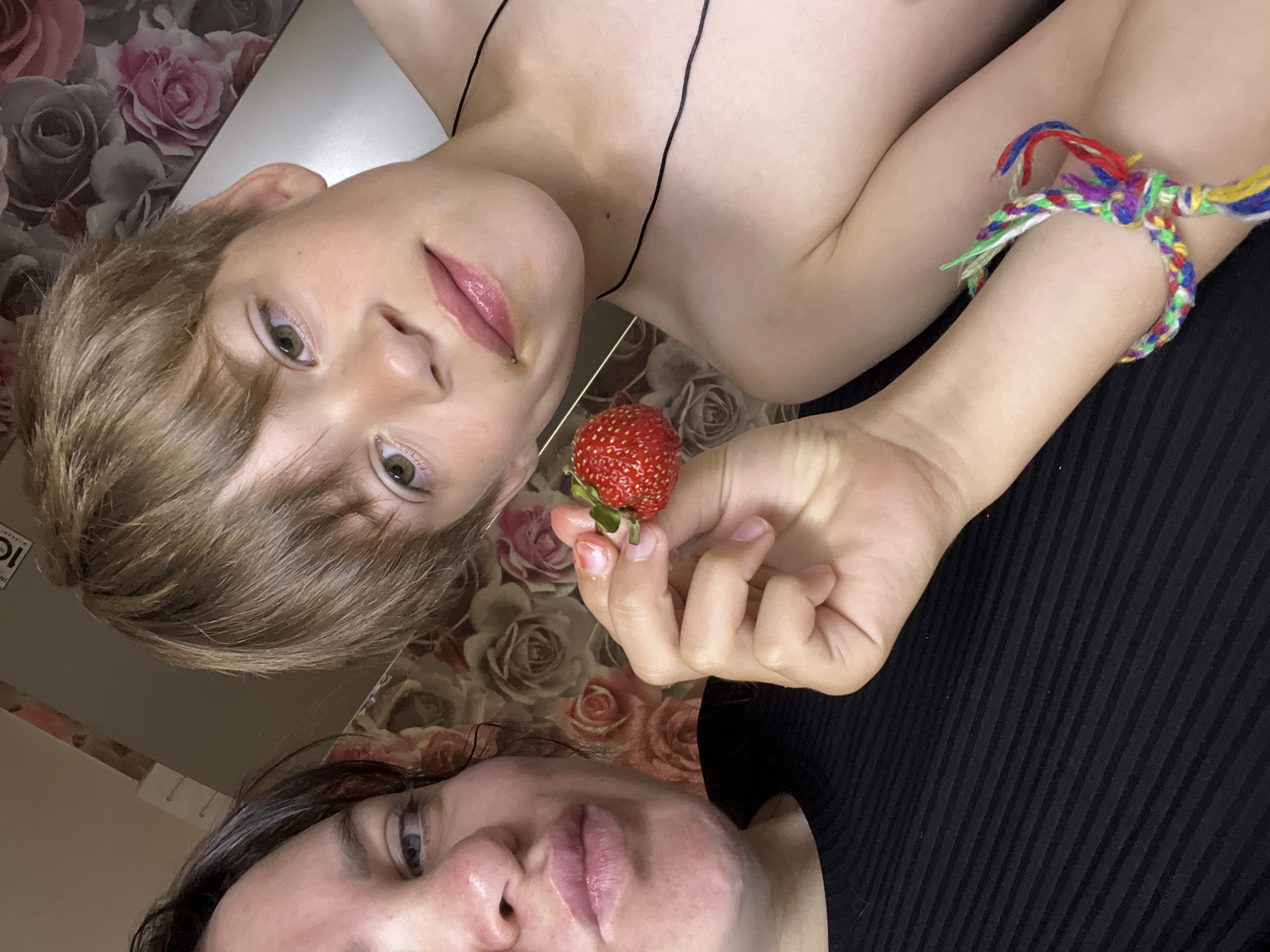 The image shows two people, a child and an adult, lying down and looking up at the camera. The child, on the left, has short blonde hair and is holding a strawberry in their right hand. They are wearing a thin black string around their neck. The adult, on the bottom, has dark hair and is wearing a black shirt. The background appears to be a surface with a floral pattern, possibly a tablecloth or wallpaper, with roses in shades of pink and grey.
The child is wearing a colorful braided bracelet on their left wrist, made of red, blue, green, white, and yellow threads. The lighting suggests it might be daytime, as there is a bright light source casting shadows. The overall mood appears casual and intimate, as if the two individuals are posing for a selfie. There is no visible text in the image. The context provided places this scene in Noviy, Russia.