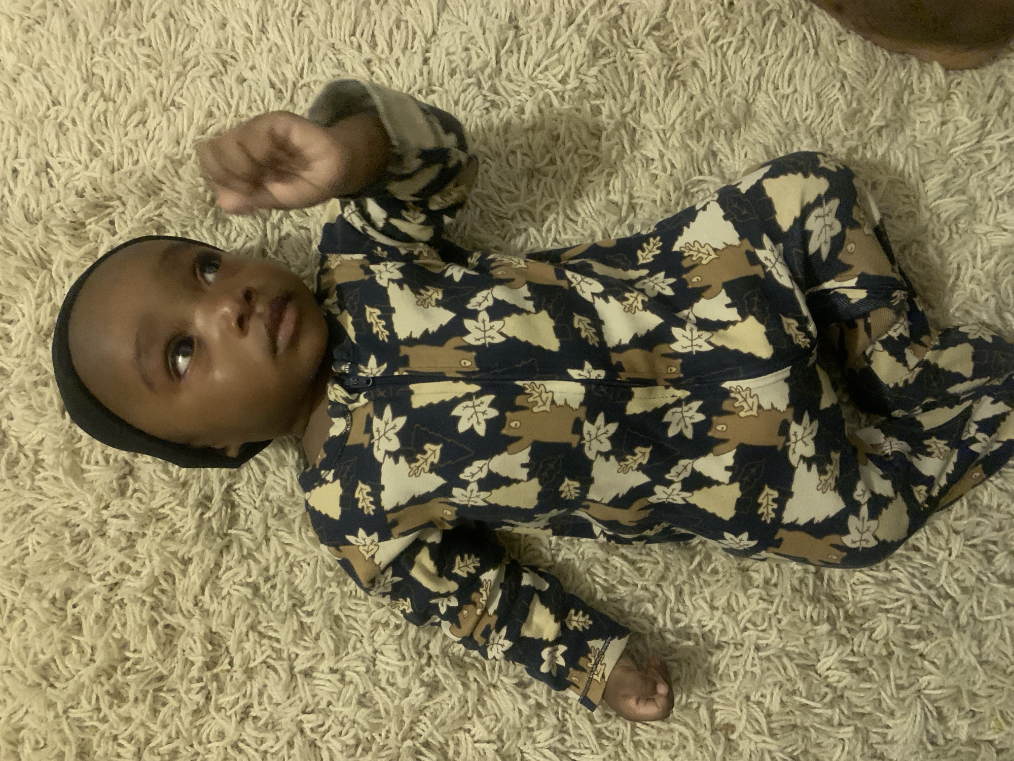 A dark-skinned infant wearing a patterned, zip-up jumpsuit and a dark-colored head covering lies supine on a light-colored, textured rug. The jumpsuit features a repeating pattern of leaves and bear figures in shades of dark blue, tan, and brown. The infant is looking upwards with their left arm raised, and their right arm resting at their side. The rug's texture appears plush and consists of tightly packed fibers. The scene is illuminated by overhead lighting. There is no evidence to suggest that the photo was taken in Barakin Akawo, Nigeria.