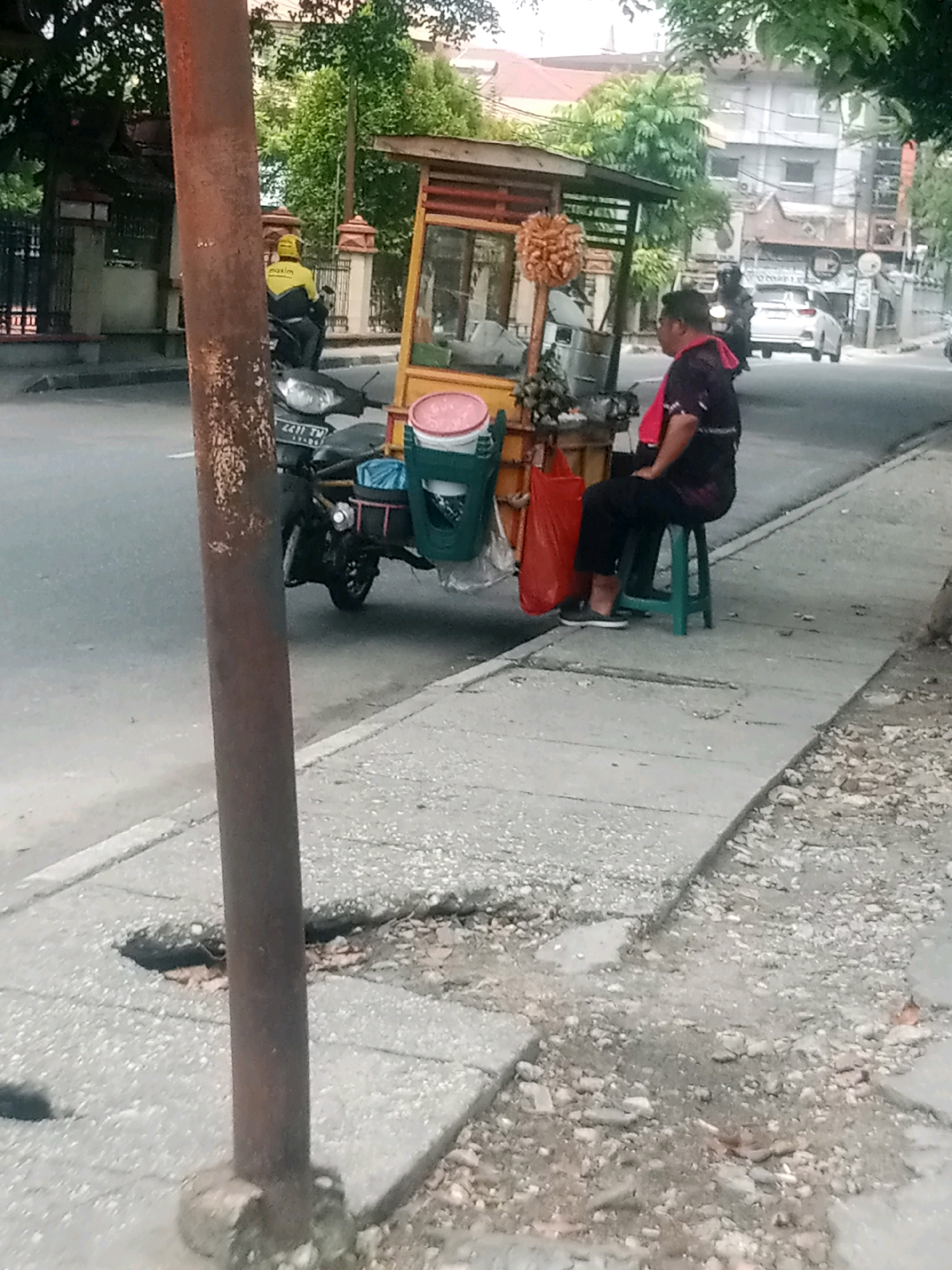 A person sits on a green stool in front of a food cart on a sidewalk in Pekanbaru, Indonesia. The person is wearing a black shirt with a pink cloth draped around their neck and black pants. The food cart is yellow and has a red awning. A motorcycle is parked behind the food cart. The sidewalk is made of concrete and is cracked in places. There is a metal pole in the foreground. In the background, there are buildings and trees. The street is paved and there is traffic.