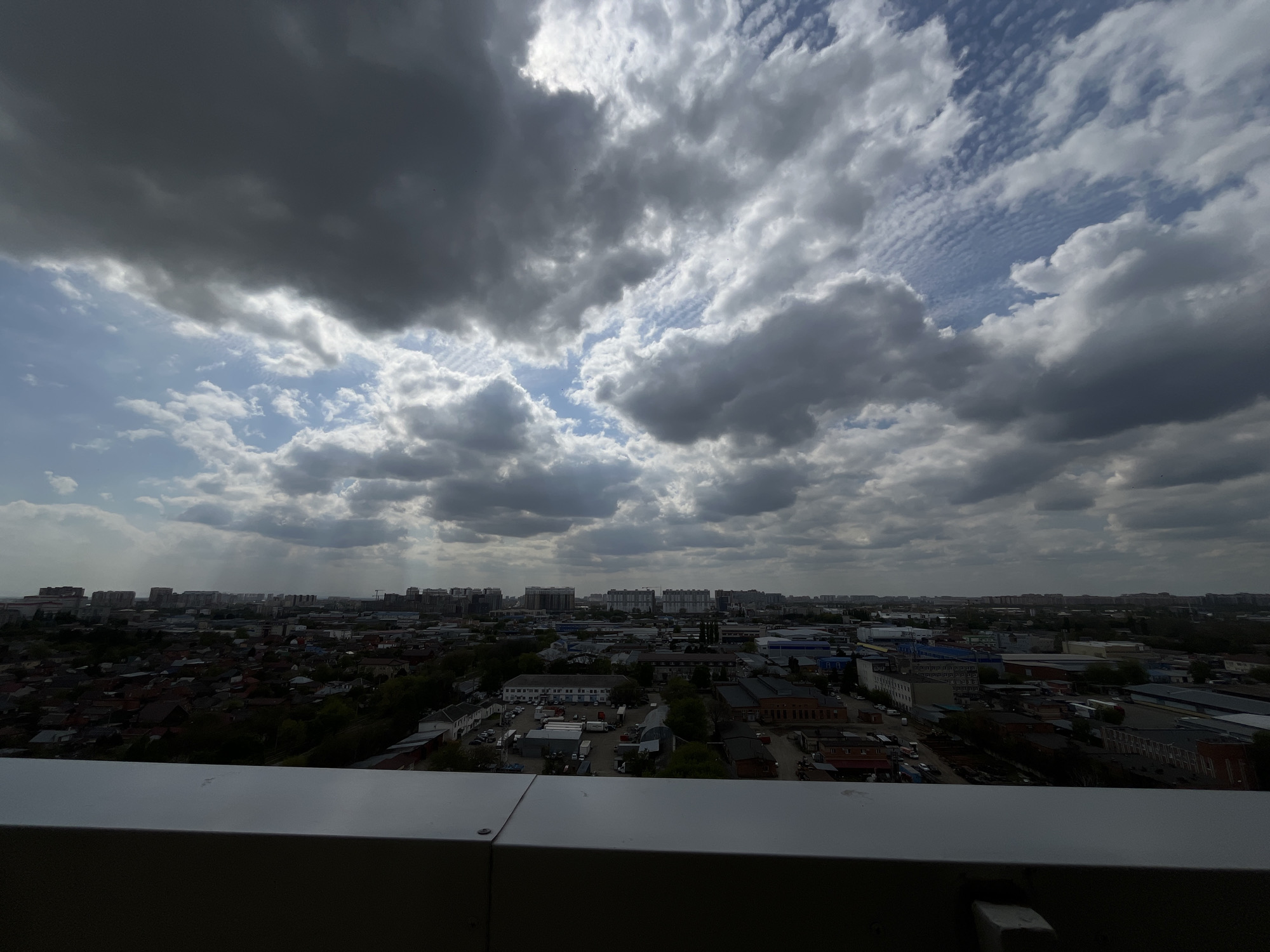 The image presents a wide, elevated view of an urban landscape under a dramatic sky, likely captured from a tall building. A white, metal railing or parapet occupies the lower foreground, indicating the high vantage point.
The sky is a prominent feature, dominated by heavy, dark grey storm clouds that cover much of the frame. Patches of brighter blue sky are visible, particularly on the left and in the upper right, where the clouds break to reveal a distinctive pattern of rippled, almost fish-scale-like clouds (possibly altocumulus undulatus). Sunlight pierces through gaps in the clouds, creating varying light and shadow effects. On the far left horizon, faint vertical streaks suggest light rain or precipitation in the distance.
Below the dynamic sky, the city sprawls into the horizon. The midground features a mix of lower-rise buildings, including residential areas with dark, often red-tiled roofs, interspersed with green trees. Further into the midground and background, there are numerous industrial or commercial complexes with flat-roofed structures, warehouses, and what appear to be storage yards and parking lots with vehicles. In the far distance, a denser skyline of taller apartment blocks and commercial buildings defines the city's edge. The overall impression is a blend of older, more suburban-like areas and modern urban development.
The scene depicts daytime conditions, but the heavy cloud cover gives it a muted, possibly pre-storm or post-rain atmosphere. There is no visible human activity or specific event taking place, and no people are discernible. No visible text is present in the image. The architectural styles and urban spread are consistent with cityscapes found in regions like Penzenskaya, Russia.