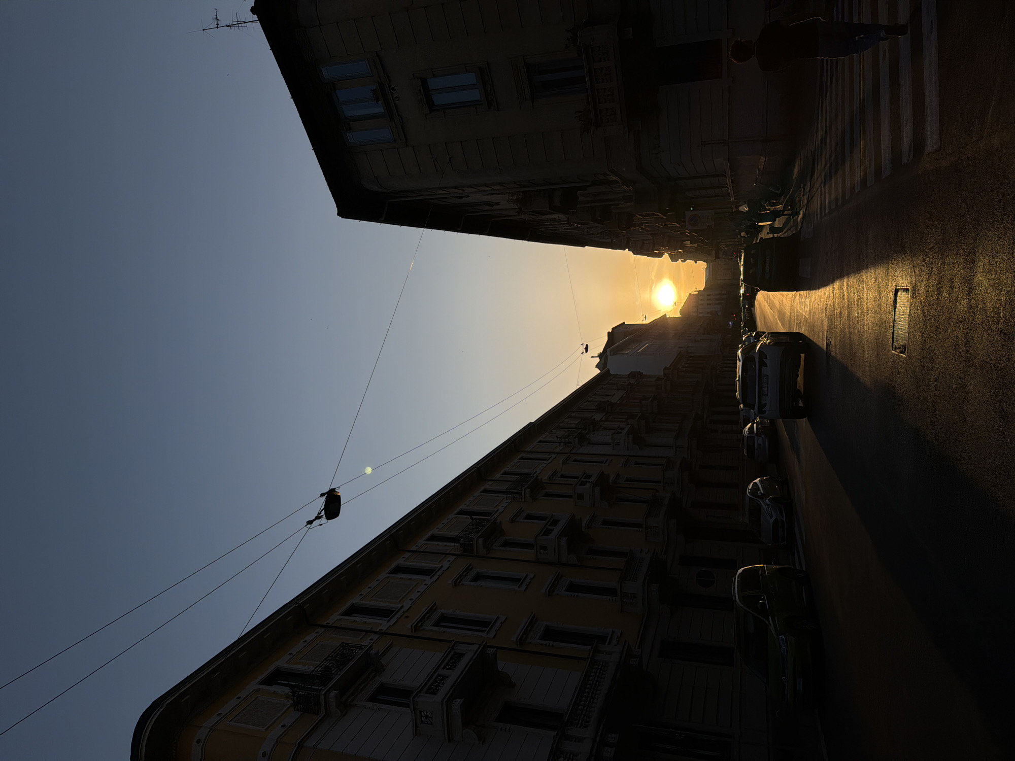 Captured at a tilted angle, the image presents an urban scene in Milan, Italy, during late afternoon as the sun approaches the horizon. Buildings line either side of the street, their detailed windows and architectural elements contrasting sharply with the shadows cast by the low sun. Parked cars are visible along the roadside, reflecting the sunlight. In the distance, the sun shines through a gap between buildings, creating a bright focal point. Overhead, power lines stretch across the sky, intersecting the buildings. A solitary figure walks across a pedestrian crossing at the end of the street, with the street illuminated by the sun.