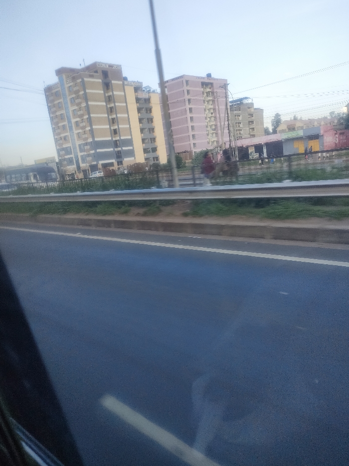 The image depicts an urban landscape in Theta ward, Kenya. In the foreground, a dark asphalt road surface with a single white lane marker is visible, suggesting it was captured from a moving vehicle. A concrete curb separates the road from a grassy median strip, which is protected by a silver metal crash barrier.
Beyond the barrier, a pedestrian area or shoulder runs parallel to the road. Several indistinct human figures are present, with one individual in red attire walking towards the right. A tall, slender utility pole extends vertically from this midground section. Behind the pedestrian area, a dark fence separates it from a series of single-story commercial buildings, some featuring yellow and orange-red facades. A white vehicle and a large dark vehicle (possibly a bus) are partially visible further back on the left.
The background is dominated by multiple multi-story residential buildings. On the left, a building identified by "WALES PLACE" signage displays a facade with alternating vertical sections of beige, light blue, and grey. To its right, a prominent pink-colored building, featuring balconies and structural elements, is marked with "JALUO HEIGHTS" and "JALUO RESIDENTS" at its apex. Both primary buildings appear to be approximately 10-12 stories high. Additional, less distinct residential structures and sparse vegetation are visible in the distant background. Overhead, multiple utility lines or power cables are stretched across the sky, which is a light blue, hazy tone indicative of daytime conditions.