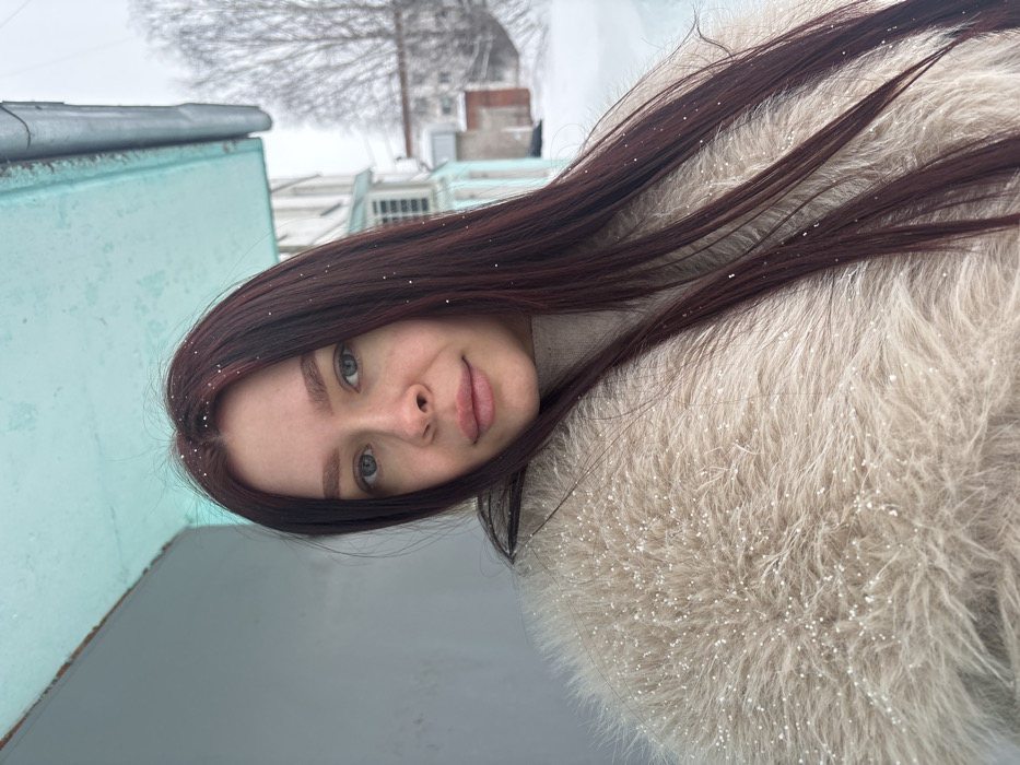 A young adult female is depicted in a close-up outdoor portrait, presumably within the city of Chaikovsky, Russia, during winter. She has fair skin, light-colored eyes, and long dark auburn hair with visible snowflakes resting on it. Her lips are full, and her expression is neutral as she looks directly at the camera. She is dressed in a light beige turtleneck beneath a voluminous, shaggy light beige faux fur coat, also dotted with snowflakes. The background to her left features a plain light gray wall topped by a bright turquoise architectural overhang with a dark gutter. To her right, a snow-covered urban landscape is visible, including several multi-story residential buildings with light-colored facades and some turquoise accents, and bare deciduous trees under an overcast sky.