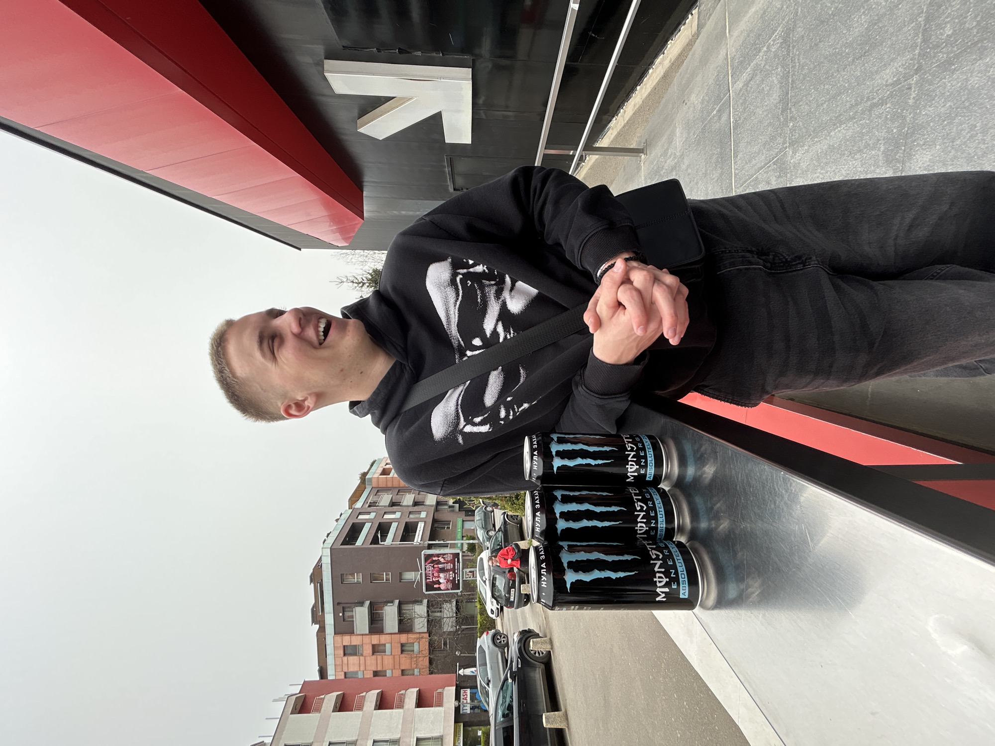 A photograph taken in Sofia, Bulgaria, depicts a fair-skinned young man with closely cropped hair and a black hooded sweatshirt with a graphic on it, wearing dark jeans and a black shoulder bag. He stands next to a silver metal surface on which four cans of Monster Energy drink are stacked, each can being the "Absolutely Zero" variety with Cyrillic text. The man has his hands clasped and is looking to his right with a smile. Behind him, there is a building with red and black structural elements and a large white arrow pointing to the right. In the background, there are more buildings, cars, and trees, suggesting an urban environment. The sky is overcast.