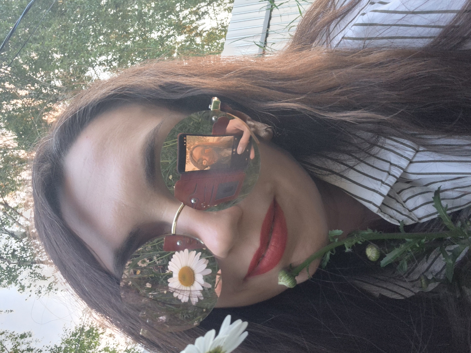 A young woman with long, brown hair is taking a selfie outdoors. She is wearing large, round sunglasses with reflective lenses and a striped shirt. In the left lens of her sunglasses, a white daisy is visible. In the right lens, her reflection is visible, holding a smartphone and taking the selfie. She has bright red lipstick on. A flowering plant is in the foreground, partially obscuring her face. The background consists of trees and a light-colored fence. The scene appears to be set in a residential area, possibly a yard or garden, in Chaikovsky, Russia.