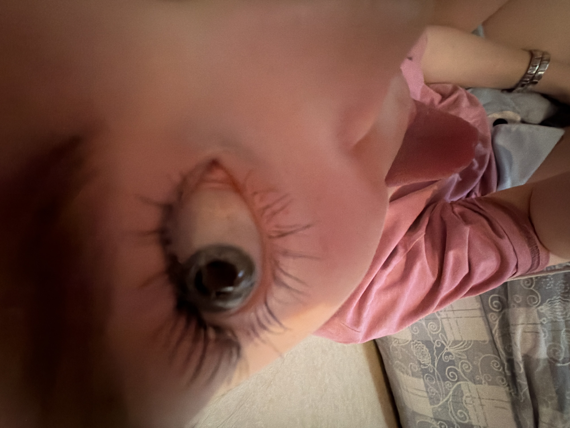 The image is an extreme close-up of a person's eye, presumably a child. The eye is brown with long eyelashes. A portion of the person's face is visible, along with a pink shirt, a metal wristwatch, and a patterned blue/gray surface that appears to be a bedspread. The image does not provide enough information to determine the location in Perm, Russia, or any actions/interactions taking place.