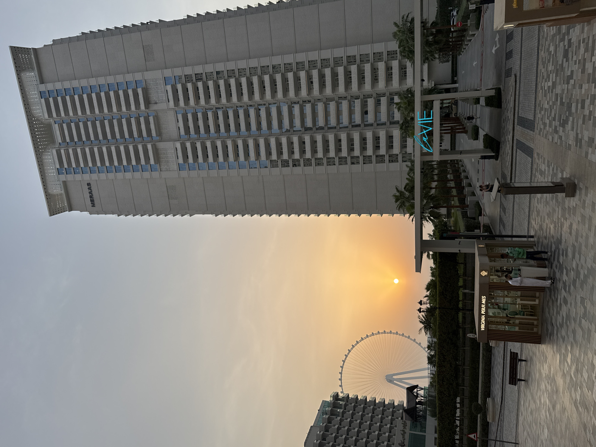 This image depicts a street scene at dusk in Dubai, United Arab Emirates, likely at the Bluewaters Island or Jumeirah Beach Residence area, given the presence of the Ain Dubai Ferris wheel and modern architecture. A tall, light-colored residential building with many balconies dominates the right side of the frame. The building has the word "MERAAS" visible near its top. To the left, a portion of another building with a similar facade and a large Ferris wheel are visible against a warm, orange-tinted sky, characteristic of sunset.
In the foreground, a small kiosk labeled "VIRGINIA PERFUMES" is situated on a paved walkway. Two men are near the kiosk; one, wearing a white thobe and ghutra, appears to be interacting with a display, while the other, in a green jacket and dark pants, stands to his left. A dark wooden bench is placed to the left of the kiosk. Further into the scene, a white archway with neon blue lettering spelling "LA VIE" is visible, with palm trees lining the path leading through it. Pedestrians are seen walking in the distance, and vehicles are present on a road beyond the archway. The ground is paved with large square tiles.
