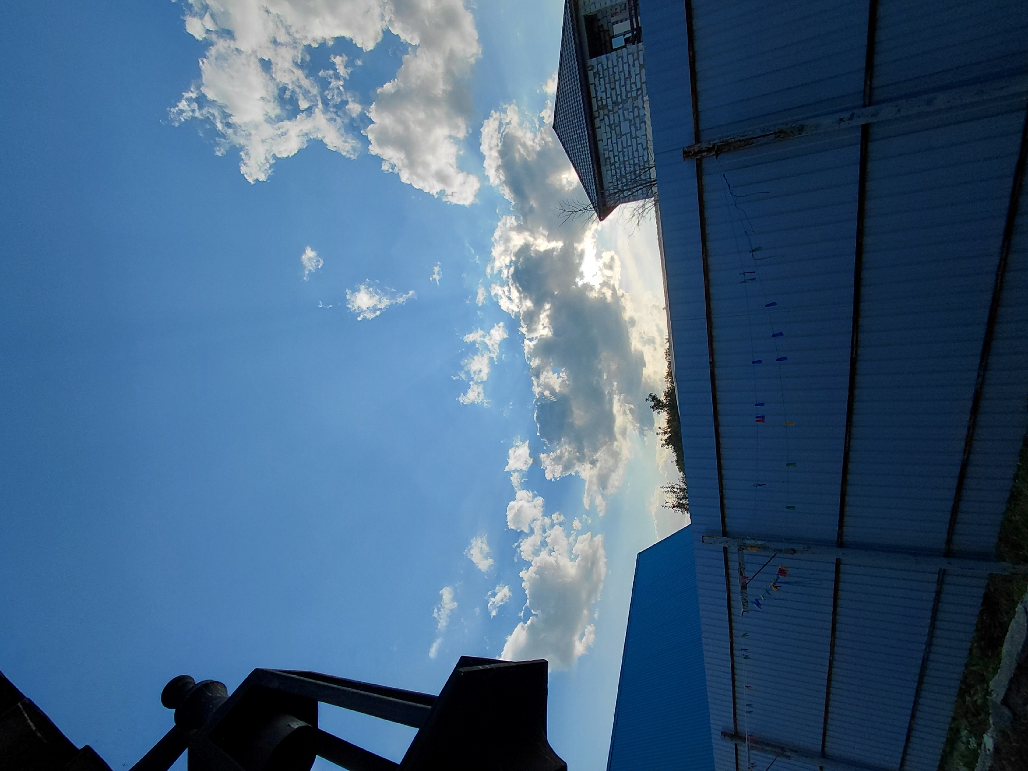 A wide-angle, low-angle, vertical shot captures a suburban scene under a bright, partly cloudy sky. The sky is a vibrant blue, with large, puffy white clouds scattered across it, some catching the sunlight. In the foreground, a dark, metallic structure, possibly a lamp post or part of a building, juts in from the left edge. Below it, the blue, corrugated metal roof of a building is visible. A tall, grey corrugated metal fence runs horizontally across the mid-ground, separating the view. Clothes pins are attached to a wire strung along the fence. Behind the fence, to the right, a house constructed from light-colored bricks with a dark tiled roof is partially visible. Trees are silhouetted against the sky in the distance. The scene appears to be in a residential area of Novosibirsk, Russia, likely during the daytime.