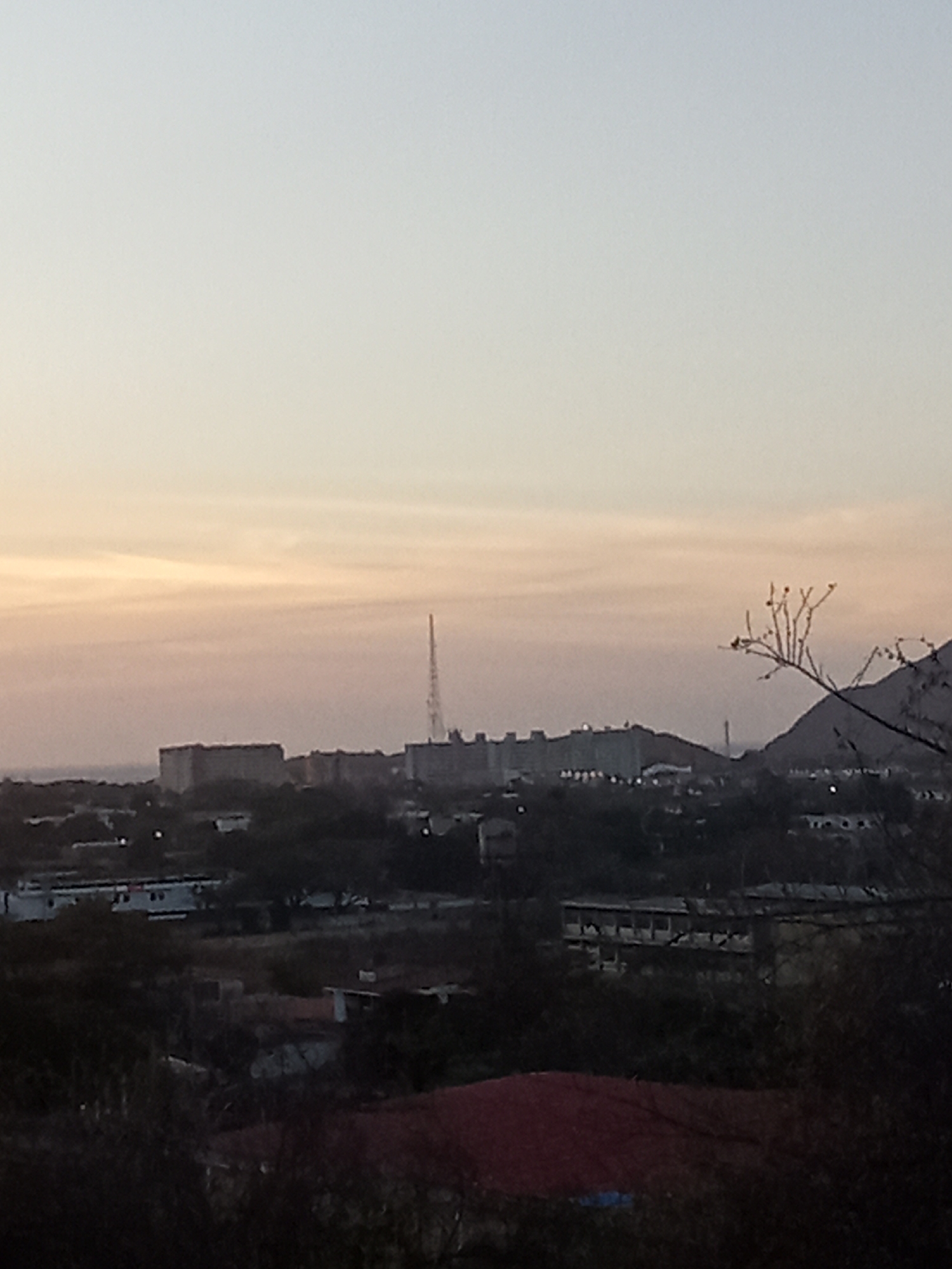 The image shows a cityscape at sunset or sunrise, possibly Juan Griego, Venezuela. There are several buildings, a communication tower, and what appears to be a low mountain or hill in the background. In the foreground, there are trees and rooftops of buildings, possibly residential areas. The lighting suggests either dusk or dawn.