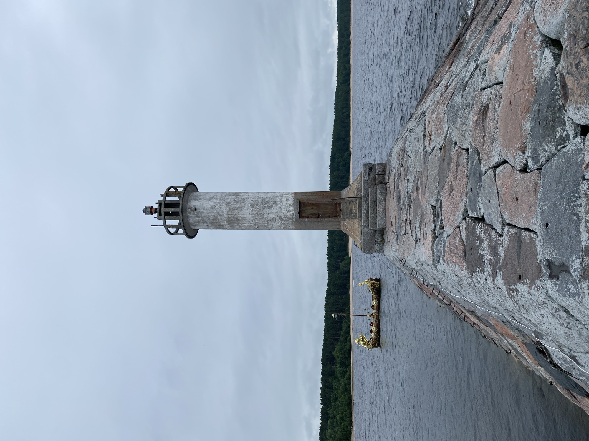 A lighthouse stands tall on a stone pier, overlooking a body of water. The lighthouse is cylindrical and weathered, with a simple wooden door at its base and a lantern room at the top. A boat with a dragon-shaped prow and stern, adorned with gold, floats in the water to the left of the pier. The background features a dense forest of green trees lining a sandy shore, under a cloudy sky. The pier itself is constructed from large, rough-hewn stones in varying shades of gray and pink. The scene appears to be set in a natural, somewhat remote location, likely Моторное, Russia, given the presence of the lighthouse, water, and forested shoreline.