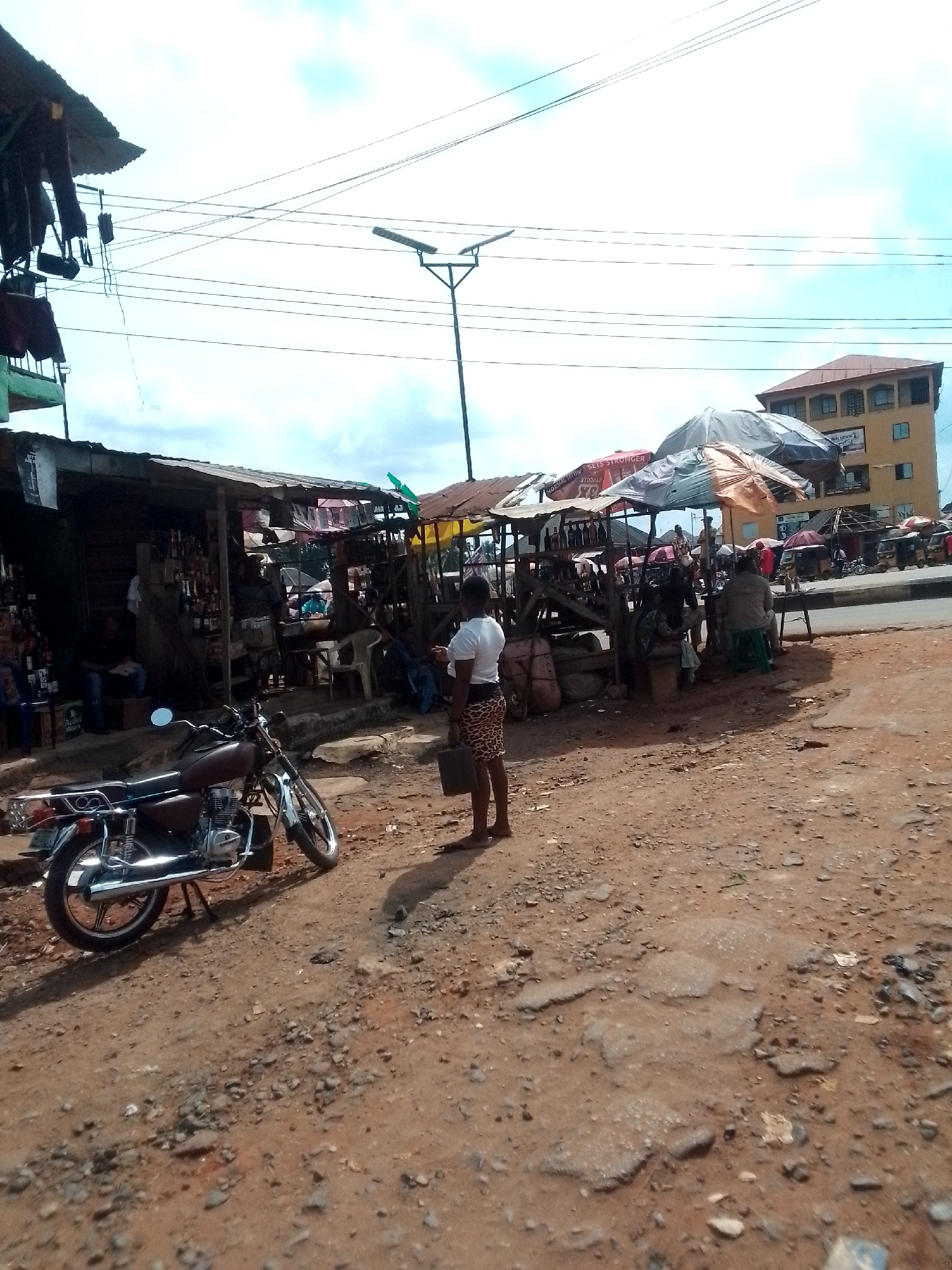 The image shows an open market scene in Orlu, Nigeria. The ground is a dusty, reddish-brown dirt, cracked and uneven. A woman stands in the foreground, wearing a white shirt and patterned skirt. A brown and silver motorcycle is parked nearby. Several makeshift stalls with corrugated iron or tarpaulin roofs are clustered together, selling goods. People are seated and standing around the stalls, engaging in commerce. A street light pole rises from the center of the market area. In the background, a multi-story building with orange roofing is visible on the other side of a street with cars on it. Overhead, electrical wires crisscross against a sky with scattered clouds.