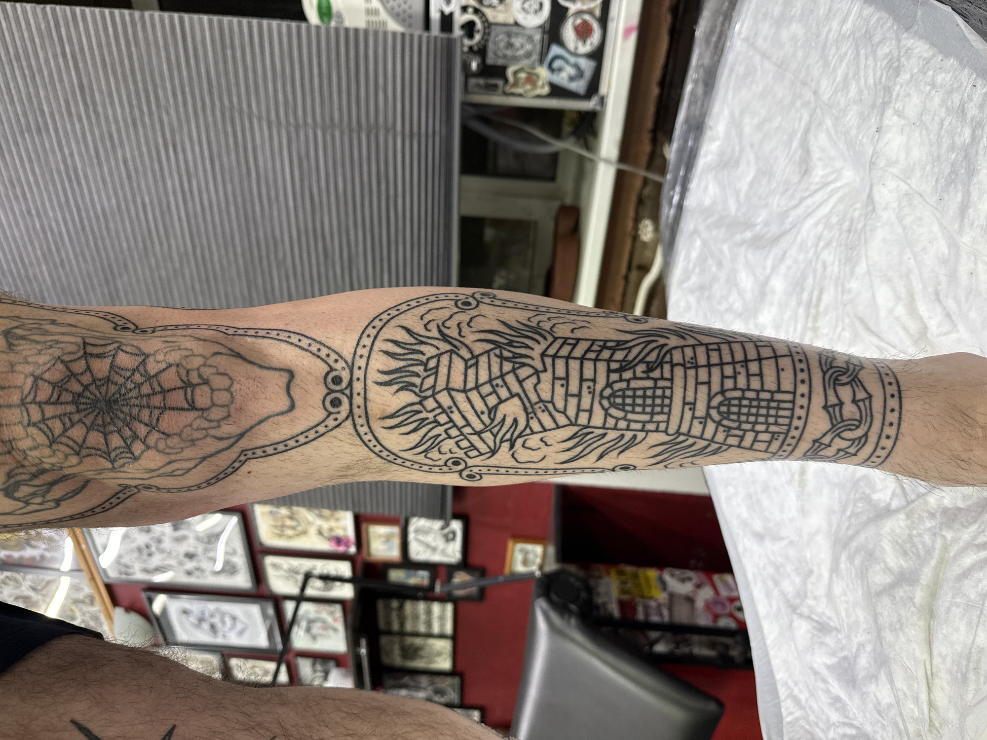 An arm with tattoos is visible in a room that appears to be a tattoo parlor. One tattoo is a spiderweb, and the other tattoo features what appears to be a building or structure. The environment includes tattoo-related artwork framed on the walls, a gray padded table, and a black stool. The location in the city of Moscow, Russia, cannot be determined from the image.