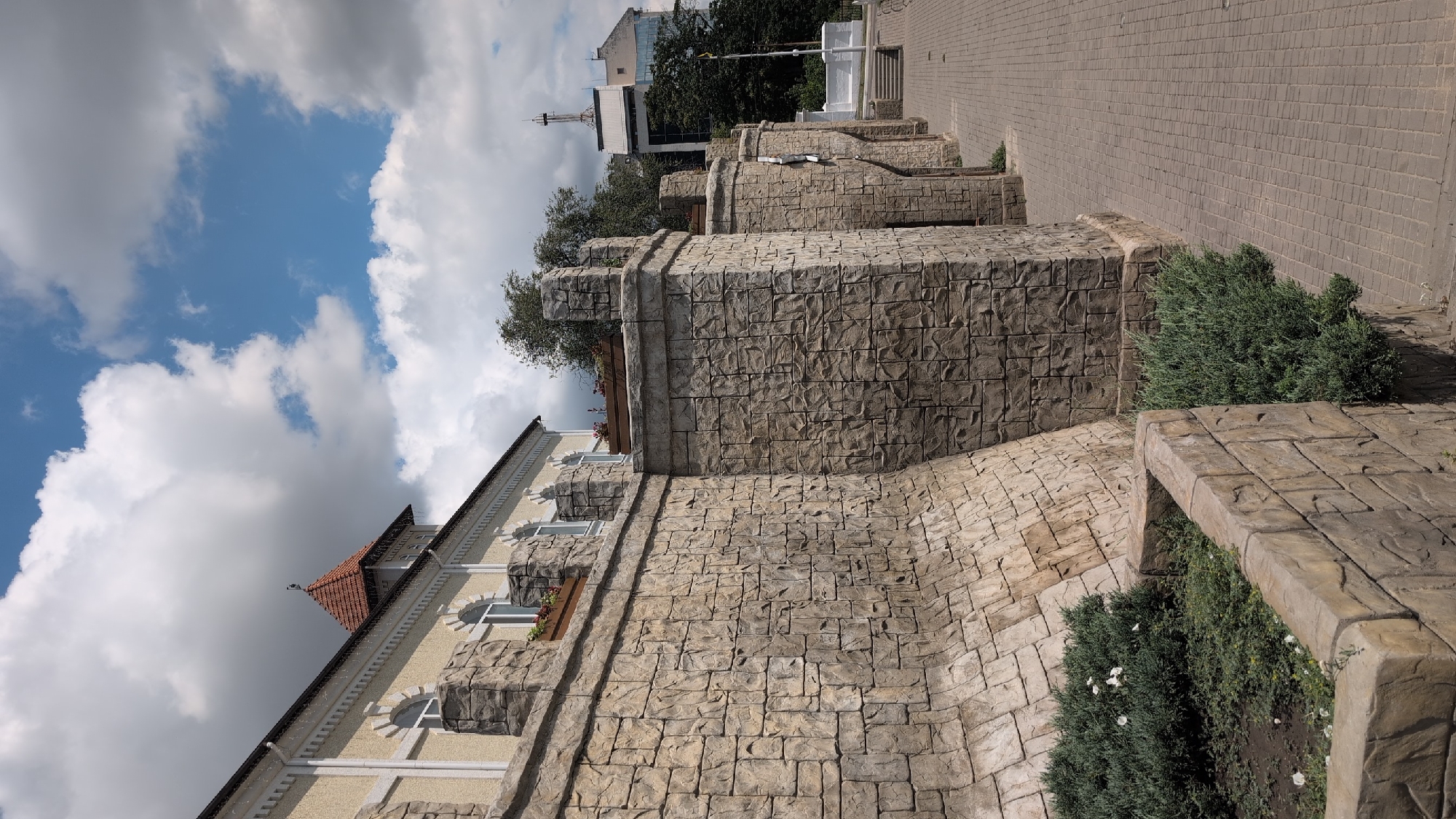 The image shows a stone wall with crenellations, reminiscent of a castle. The wall is made of irregularly shaped, textured stones in shades of beige and brown. A building with a pale yellow facade and arched windows is visible behind the wall. The building has a red-tiled, conical roof on one section. A paved walkway runs alongside the wall, made of grey square cobblestones. Green bushes are planted in front of the wall, some with small white flowers. In the background, other buildings and trees can be seen, under a partly cloudy blue sky. The scene depicts an outdoor architectural feature in a city setting.
