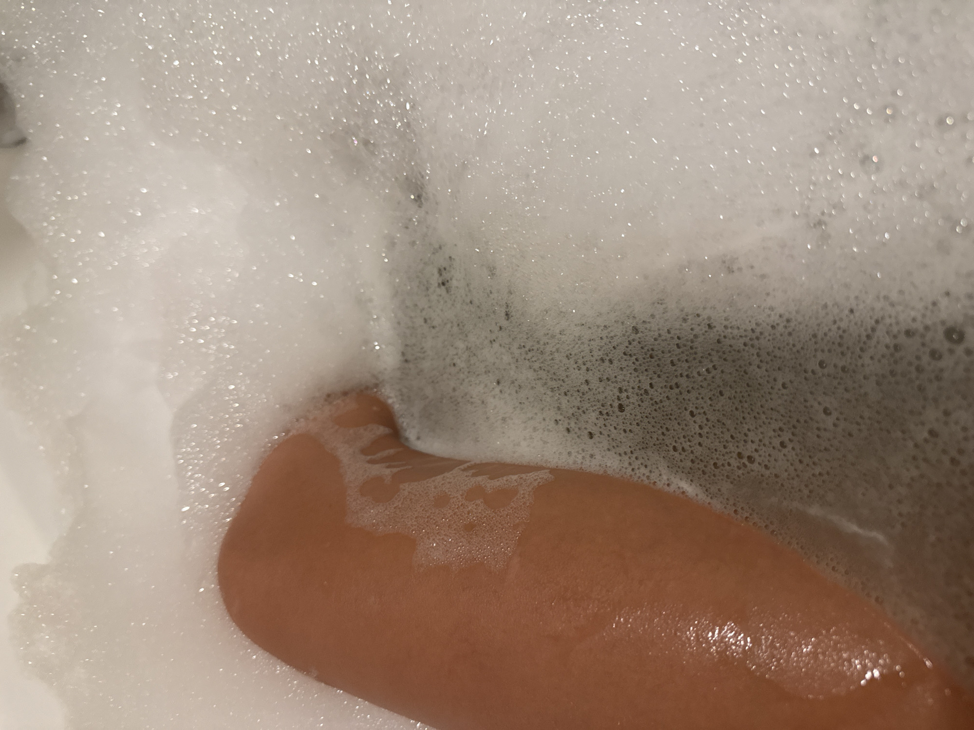 The image shows a close-up view of a person's leg submerged in a bathtub filled with white foam. The leg is smooth and appears to be in warm water, indicated by the way the light reflects off the wet skin. The foam is bubbly and fills most of the frame, obscuring what lies beneath the surface. The scene is indoors, likely in a bathroom. There is no information in the image to suggest a specific location in Chișinău, Moldova.
