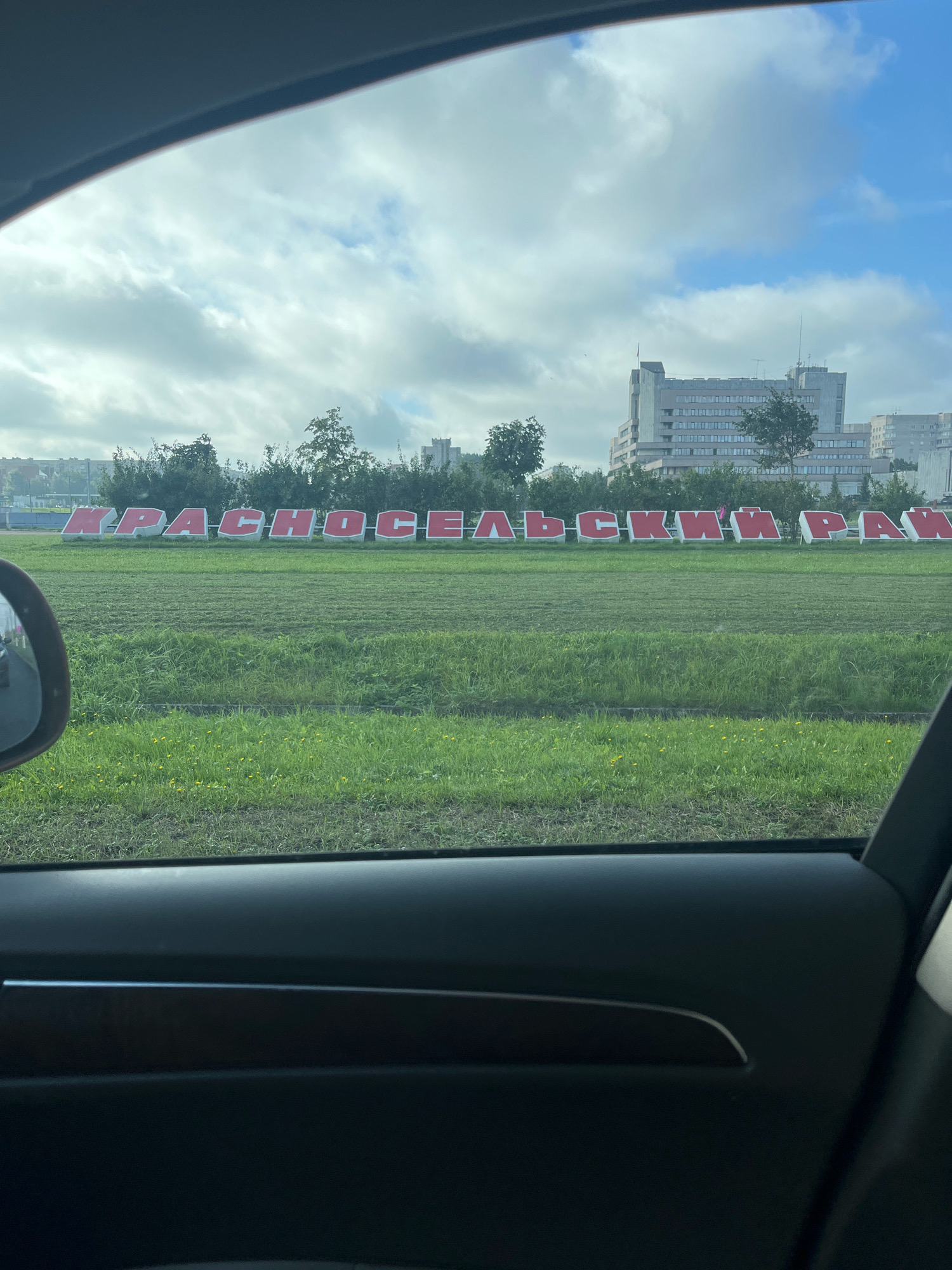 The image shows a view from inside a car, looking out a window at a landscape with large red letters spelling out "КРАСНОСЕЛЬСКИЙ РАЙОН" (Krasnoselsky District) in front of a line of trees and buildings. The letters are on a grassy field. In the background, there are several multi-story buildings, some appearing to be residential or office buildings, under a cloudy sky. The foreground shows the interior of the car, including a portion of the car's door panel and a rearview mirror reflecting the scene outside. The overall impression is of a roadside view in a suburban or semi-urban area of Saint Petersburg, Russia.