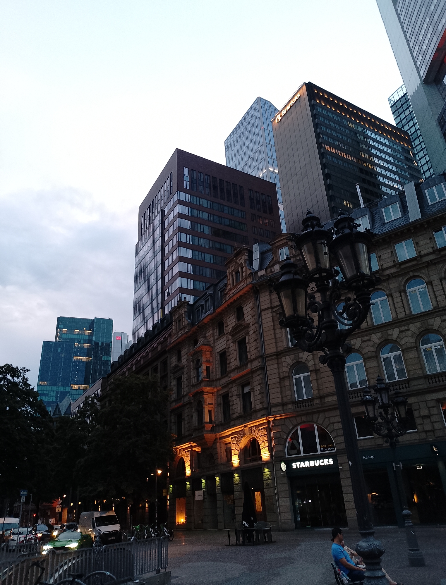 The image shows a street view in a city with modern and historic buildings. In the foreground, an ornate lamppost stands on a paved area. To the right of the lamppost, a Starbucks store is visible with its logo lit up. Next to Starbucks, an Aesop store is also present. A man is seated on a bench in front of the Starbucks. In the background, several tall, contemporary skyscrapers are visible, some with illuminated windows. The sky above is overcast. To the left, there is a street with vehicles, including a white van and a green car. Trees are also present along the street. The architecture suggests a European city setting.