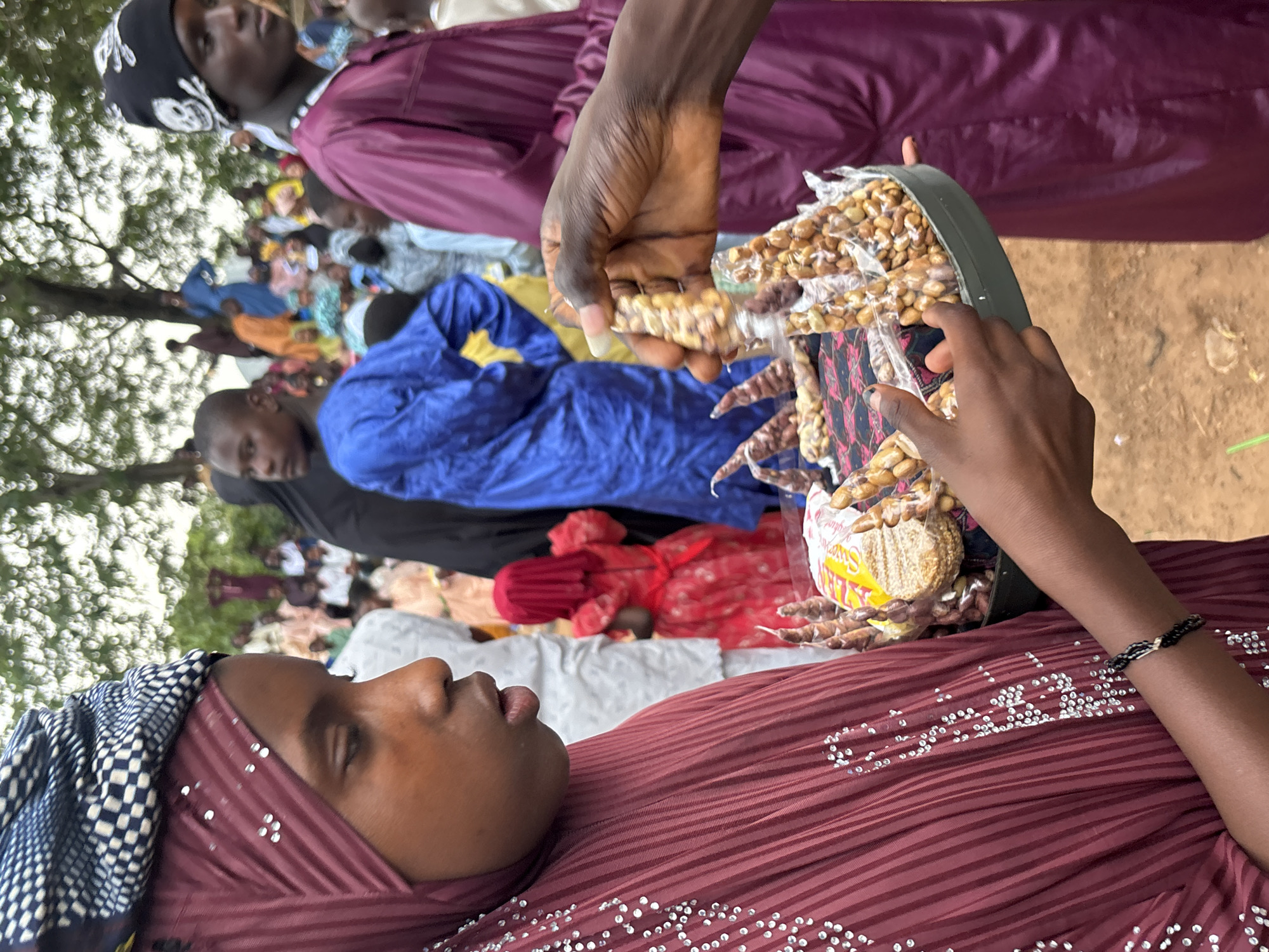 In Jos, Nigeria, a young girl in a burgundy hijab adorned with white beads is holding a circular tray filled with packaged snacks. The tray also contains a small, circular, light-colored item wrapped in yellow and white packaging. Her left hand is extended, offering a packaged snack that appears to be a string of nuts. Her right hand rests on the tray. Behind her, a crowd of people is visible, many dressed in traditional attire. To her right, a man in a purple robe is standing, his left hand also extended, possibly reaching for a snack. The environment appears to be an outdoor gathering, possibly a market or a festival, with trees and a dusty ground visible in the background.