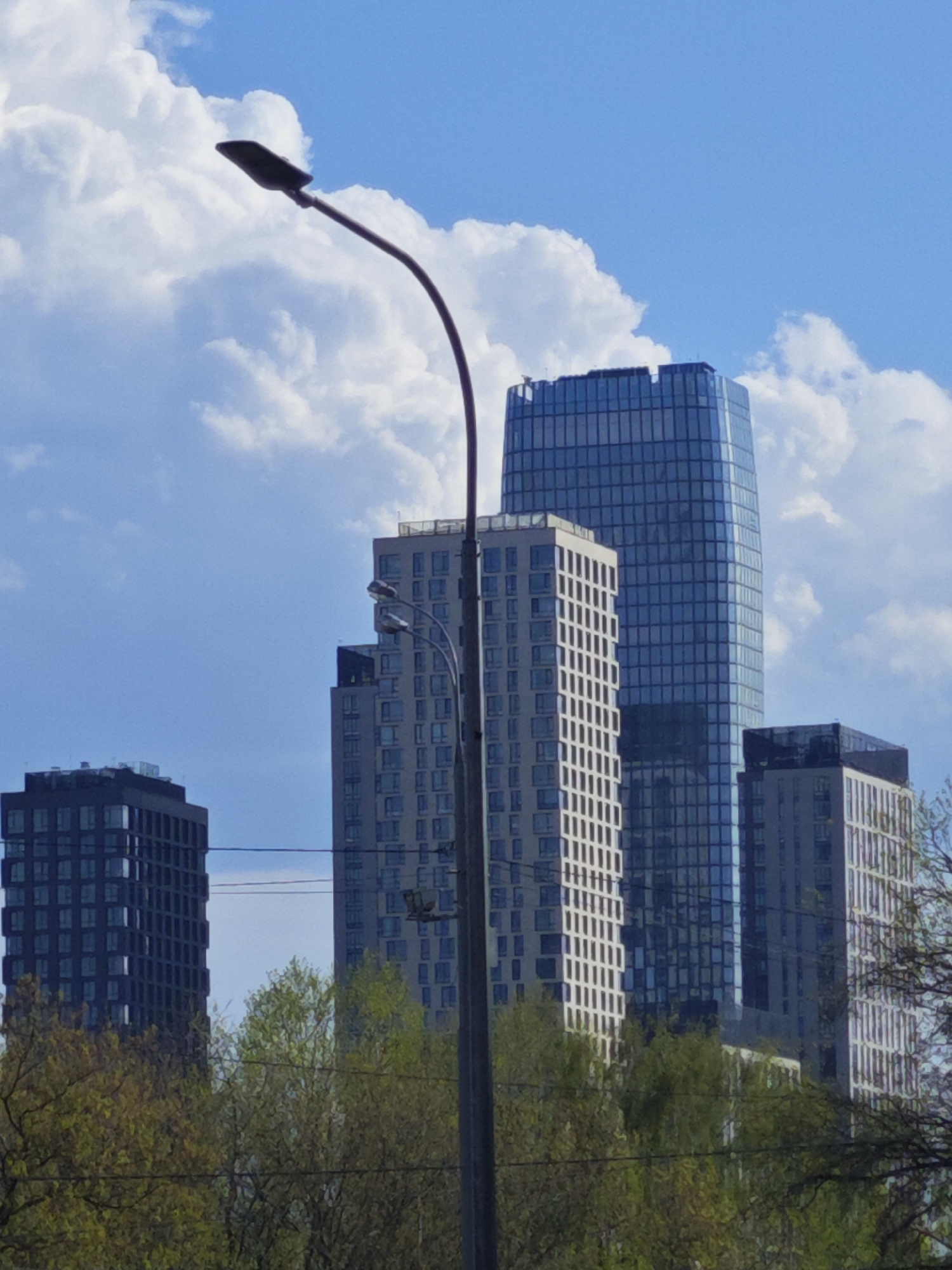 The image shows a cityscape in Moscow, Russia, featuring four high-rise buildings of modern architectural design. The tallest building is clad in reflective blue glass, flanked by two buildings with white facades and gridded window patterns. A darker, shorter building is visible to the left. In the foreground, trees with green foliage partially obscure the lower portions of the buildings. A street lamp with a curved arm dominates the center-left of the frame. The sky is blue with cumulus clouds.