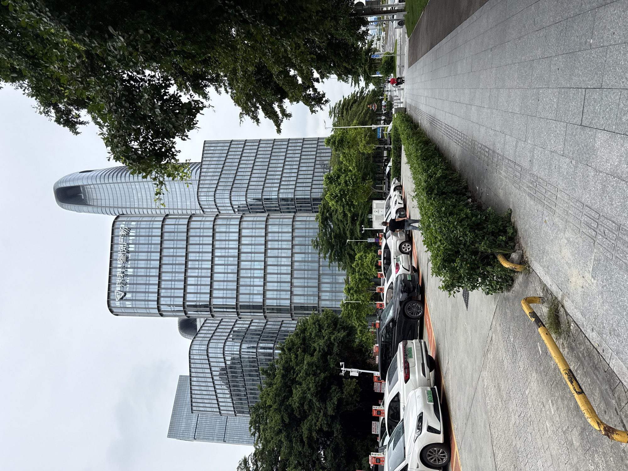 This is a medium-shot, eye-level, outdoor, vertical photograph taken in Haizhu District, China. The image shows a row of modern, glass-facade office buildings with distinct architectural styles. In the foreground, a line of parked cars, predominantly white and one black SUV, are visible in a parking lot. A person in a black t-shirt and jeans stands near the cars, facing away from the camera. Adjacent to the parking lot is a paved walkway with a low, green hedge bordering it. Further down the walkway, a person in a red top rides a scooter. The background features lush green trees and a cloudy sky. The scene appears to be a business or commercial district.