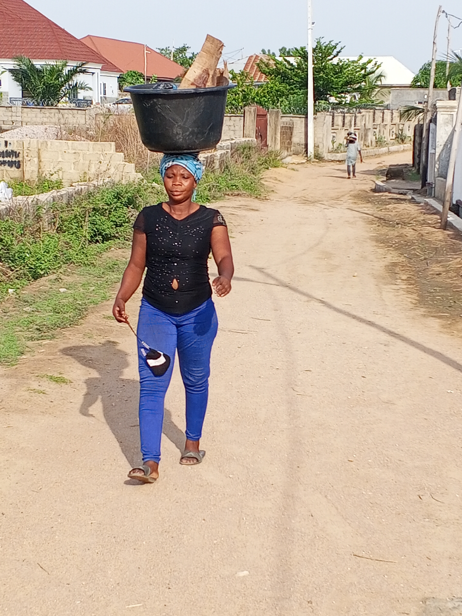 A woman walks along a sun-drenched, unpaved road in Adonkolo, Nigeria, expertly balancing a large, black plastic basin filled with pieces of wood or similar material on her head. She is positioned in the foreground, moving towards the left side of the frame. She wears a light blue patterned headwrap, a short-sleeved black top with some distressed details, and form-fitting blue jeans. On her feet are open-toed sandals. In her right hand, she holds a dark object, possibly a folded face mask attached to a string or strap.
The road is made of light brown dirt or sand, showing clear shadows cast by the subjects, indicating bright midday or early afternoon sunlight. Along the left side of the road, there's a low concrete block wall partially covered by green weeds and sparse vegetation. In the middle ground, residential buildings with reddish-brown tiled roofs are visible behind more foliage and fences.
Further down the dusty road in the background, another individual is seen walking away, also appearing to carry something on their head. A white utility pole stands tall on the right side of the road, and more concrete block fences line the right side of the scene. The overall atmosphere depicts daily life and labor in a semi-urban or rural Nigerian setting under clear skies. No visible text is present in the image.