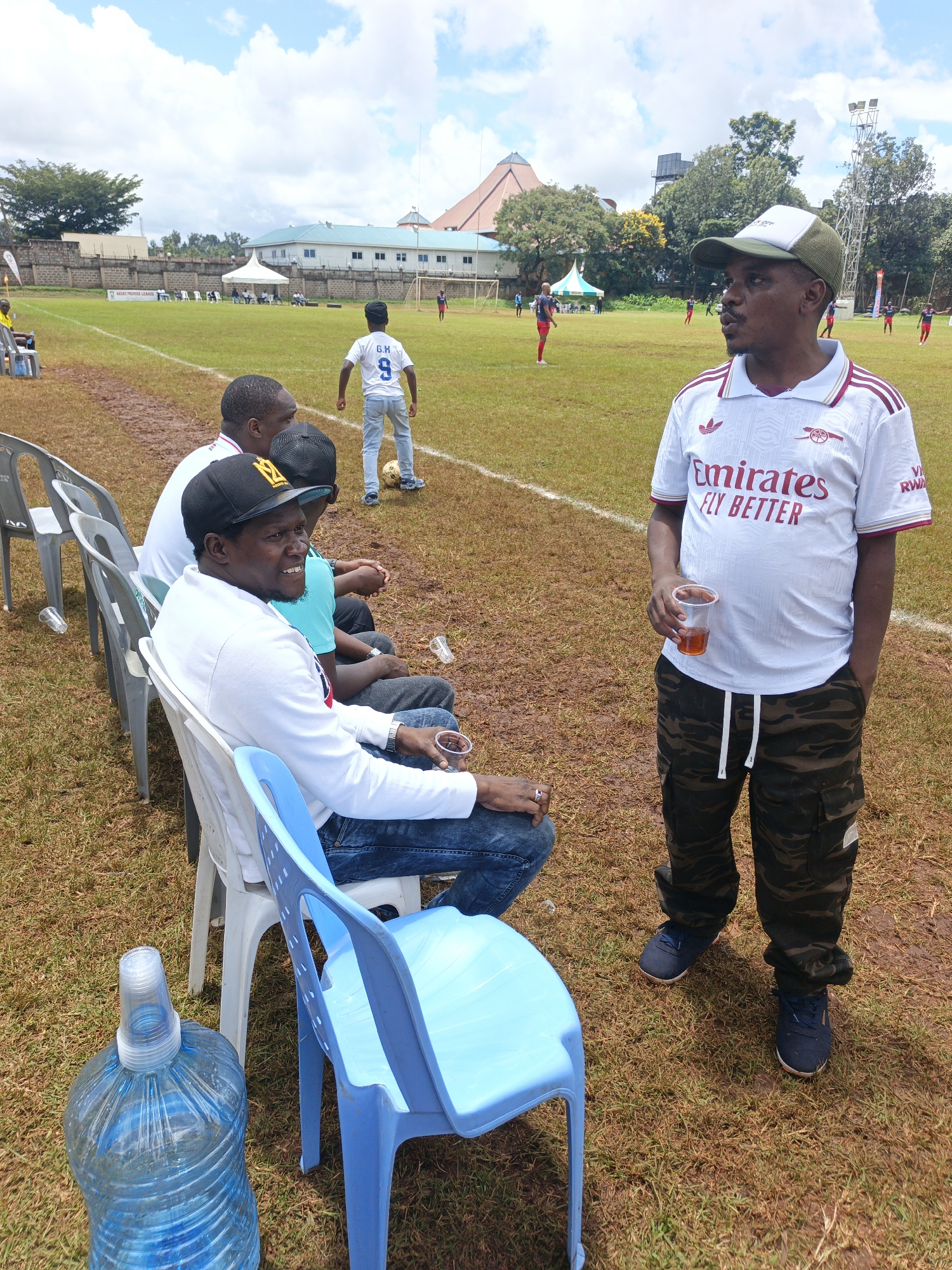 The image presents a scene on a grass field, potentially a soccer field, in Nairobi, Kenya. Several individuals are present. In the foreground, three men are seated on plastic chairs. The man closest to the camera wears a white sweater, blue jeans, and a black cap with gold lettering, and holds a plastic cup with a dark liquid. Next to the chairs is a large bottle of blue water with a plastic cup on top. Standing in the foreground is another man wearing a white Adidas jersey with "Emirates Fly Better" printed on it, camouflage pants, a cap, and holding a plastic cup. In the background, a boy wearing a jersey with the number 9 and the initials "G.M." stands near the center of the field. Several other people appear to be playing soccer. The environment includes buildings, tents, a soccer field, and trees. The lighting suggests it is daytime under a partly cloudy sky. The field appears muddy in places.