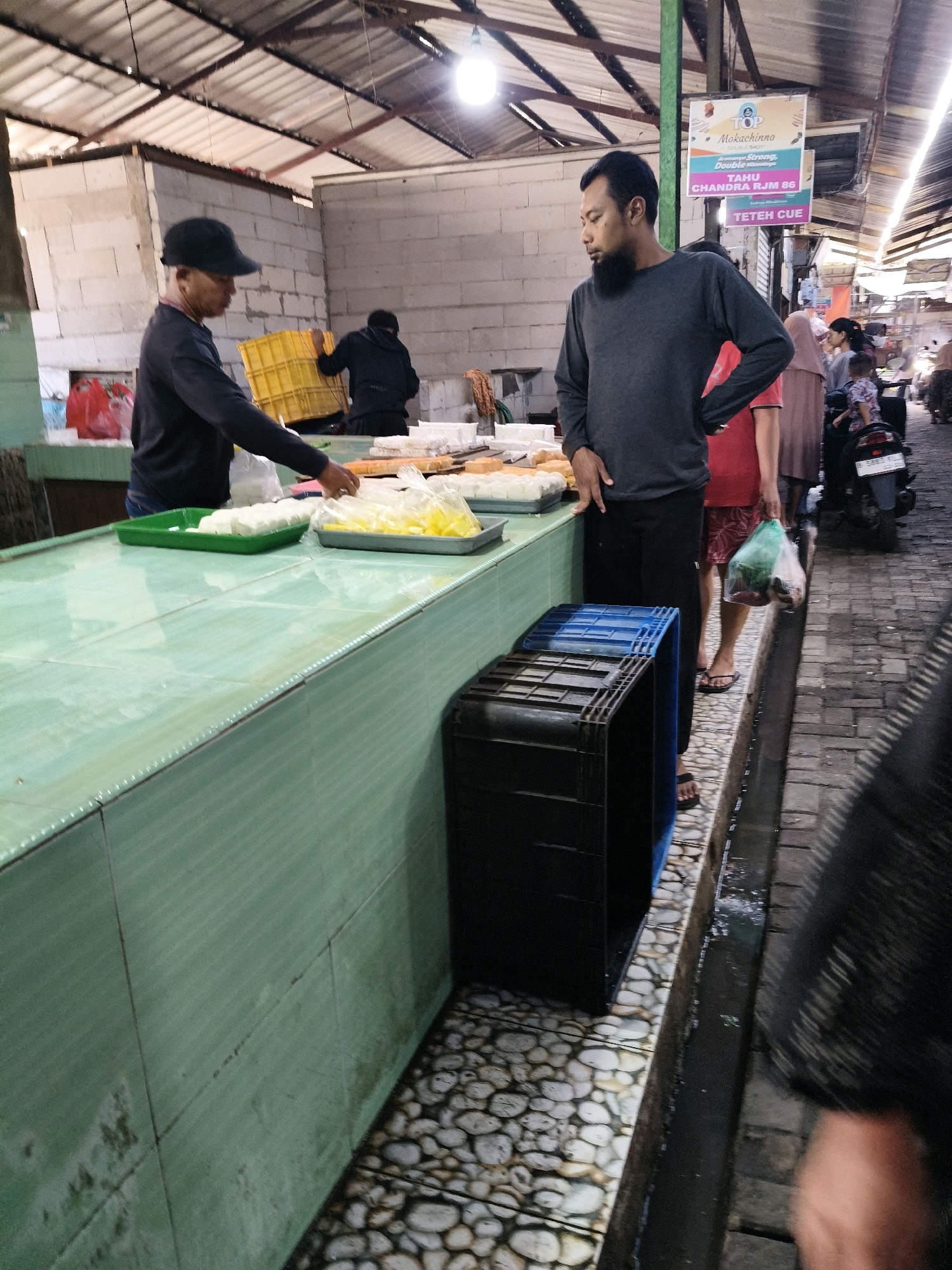In Setiamekar, Indonesia, a market scene unfolds under a corrugated metal roof, illuminated by overhead lights. Two men stand at a green-tiled counter laden with food items, one a vendor wearing a dark long-sleeved shirt and a black baseball cap, the other a customer in a dark grey long-sleeved shirt and black pants, sporting a beard. The vendor is attending to a tray of white, rectangular food items, possibly tofu or cheese, while the customer observes. Behind them, a man in a black hoodie carries a yellow basket. The counter’s surface is tiled and worn. In the foreground, two black plastic crates are stacked. The ground is a mosaic of light-colored pebbles. To the right, a motorcycle is parked near a cluster of people, including a woman in a pink headscarf and a child. Signs advertising food products are visible on a pole. The atmosphere suggests a bustling local market.