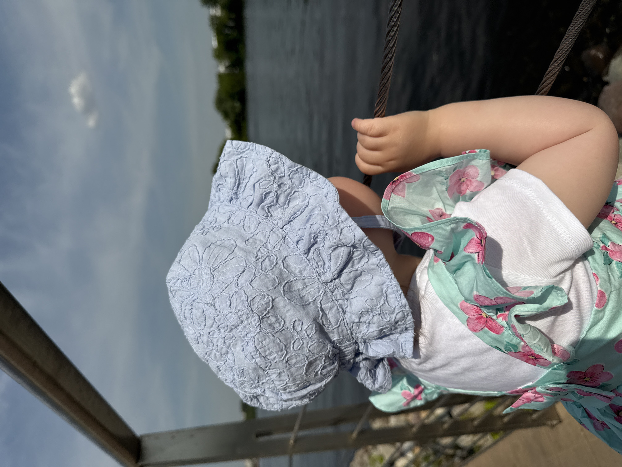 A young child, observed from a rear-side perspective, stands outdoors on what appears to be a waterfront embankment in Moscow, Russia. The child wears a light blue sun hat with intricate embroidered floral patterns and a light teal dress featuring pink floral designs, worn over a white short-sleeved top. The child's right hand is clasped around a thick, braided metal cable that forms part of a railing system. The child gazes towards a dark body of water in the mid-ground, beyond which green trees and indistinct urban structures are visible on the distant bank. The sky above is partly cloudy, with a single detached cumulus cloud visible in the upper right. A metal structural element, possibly part of the railing, is present in the upper left background. The scene is illuminated by natural daylight.
