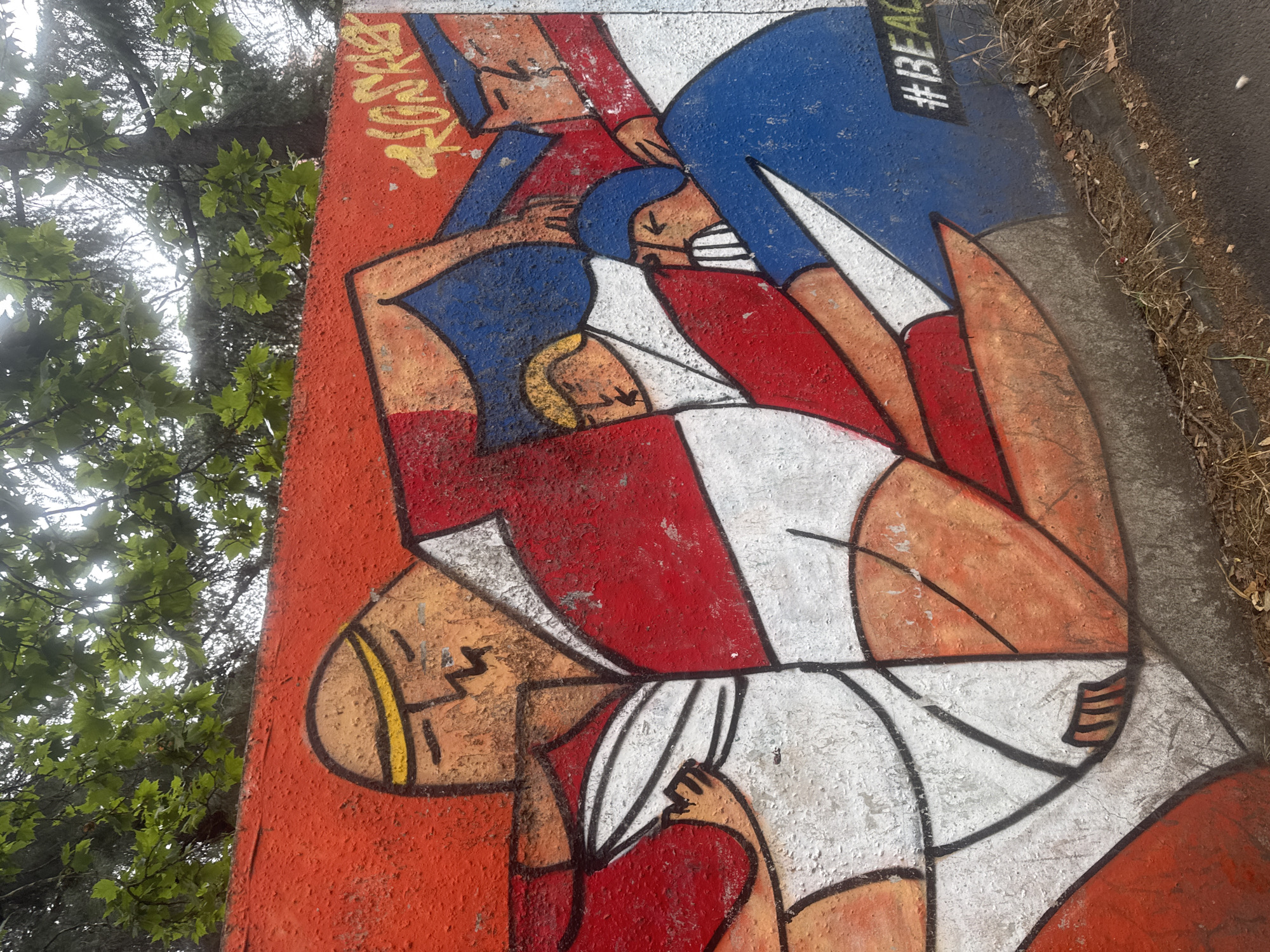 This is a photograph of a mural painted on a concrete wall. The mural depicts several stylized figures in red, white, and blue. One large figure in the foreground has a yellow band around its head and is wearing a white garment with red accents. Other figures are smaller and rendered in blue and red. There are also some yellow graffiti tags on the upper right of the mural, including what appears to be "ROSKIE." In the lower right corner, the hashtag "#BEAC" is visible in white lettering on a black rectangle. The wall is situated outdoors, with green foliage from trees visible above it. The ground in front of the wall is covered with dirt, dry grass, and leaves, with a paved surface to the right. The mural is located on the embankment of the Mtkvari River in Tbilisi, Georgia, near the Bridge of Peace.