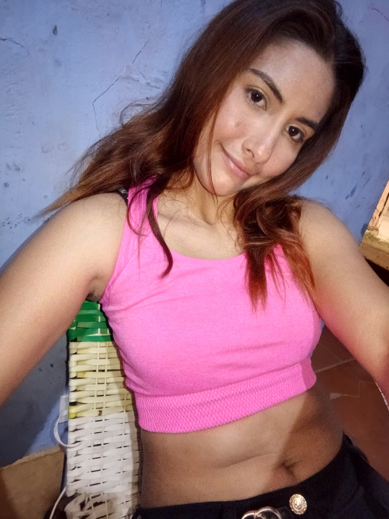 The image shows a young woman with long, brown hair, wearing a pink tank top and black pants. She is seated in front of a blue, textured wall. To her left, a portion of a white and green woven chair is visible. Her body is angled towards the camera, and she is looking directly at the lens with a slight smile. The lighting appears to be artificial, casting soft shadows on her face and skin. The background is simple and uncluttered, suggesting an indoor setting. There is no indication of the specific location within Juan Griego, Venezuela.