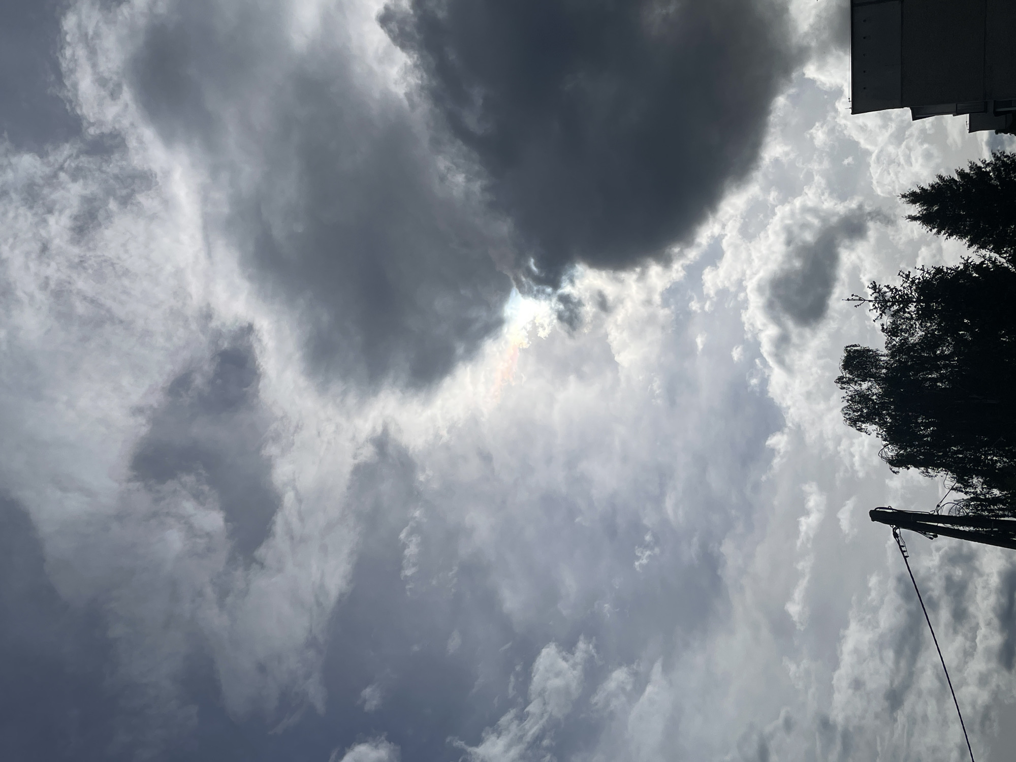 The image shows a sky with a mix of dark and light clouds. The sun is partially obscured by the clouds, creating a bright spot. In the upper right corner, there is a silhouette of a building and trees. A utility pole extends into the sky from the lower right. The location is identified as a city of Unknown, Unknown.