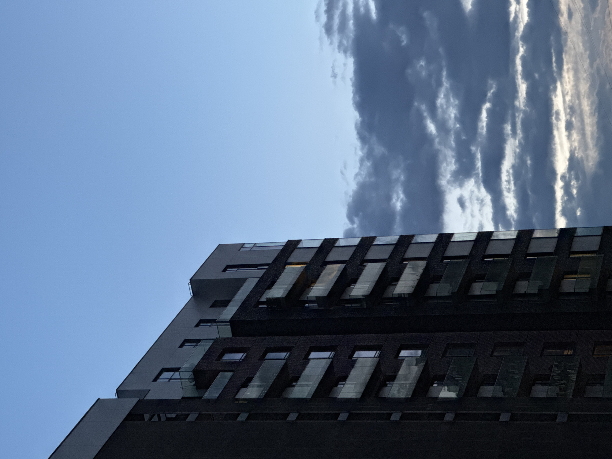 A contemporary, dark-colored building stands prominently against a dynamic sky, viewed from a low angle. The image is composed vertically, with the building occupying the lower right and central portions of the frame.
The **building** is a multi-story structure characterized by a modern, geometric design. Its facade is predominantly a very dark grey or black, with some lighter grey sections contributing to its angular profile. Numerous rectangular windows are visible, and distinctive glass-fronted elements, possibly balconies or architectural sunshades, protrude at an angle, reflecting ambient light. The building's sharp lines and dark material provide a stark visual contrast with the sky.
The **sky** fills the upper and left areas of the photograph. The upper left corner features a clear, bright blue sky. Moving towards the right and center, a mass of dark, textured clouds dominates, with some areas appearing brighter due to sunlight hitting their edges, creating streaks of white and light grey. This suggests a partially cloudy day.
There are no **people** or specific **activities** taking place; the image is an architectural study.
**Notable details** include the strong contrast between the clear blue sky and the dramatic, dark clouds, as well as the angular, modern architecture of the building. The lighting, with its bright sky and illuminated cloud edges, suggests it could be mid-day or late afternoon. The overall impression is one of urban modernity. Given the provided context, the building is located in Moscow, Russia, though no specific city landmarks are visible to confirm this visually. No text is visible in the image.