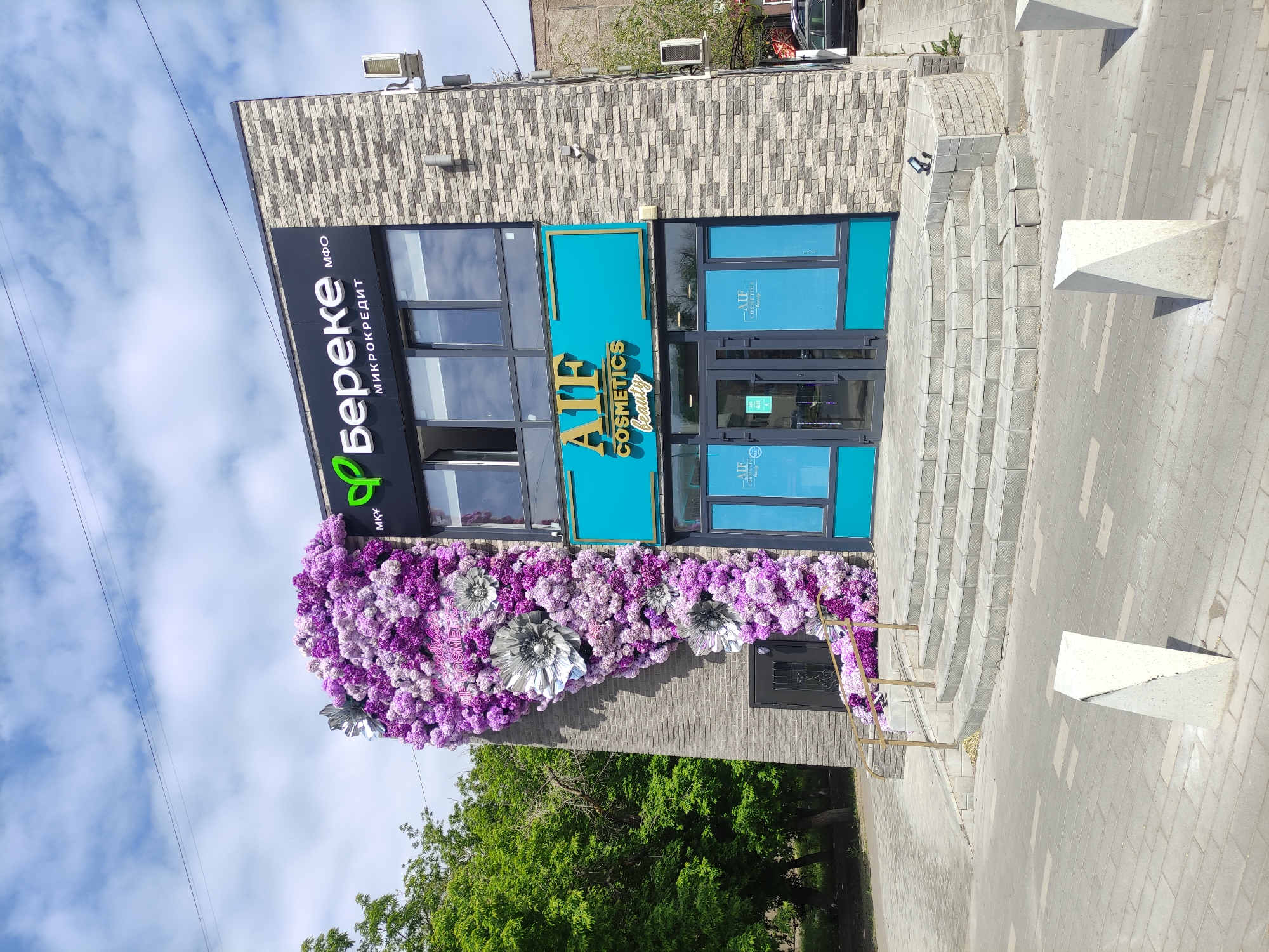 The image depicts a building facade in Pavlodar, Kazakhstan. The building appears to have two distinct businesses: one is a microcredit organization, identified by a sign in Cyrillic script that reads "Береке МКК МФО" (Bereke MKK MFO). The other business is a cosmetics store named "AIF Cosmetics Beauty."
The facade of the building is decorated with a dense arrangement of purple and white flowers, partially obscuring the lower section. The building is gray with a brick-like pattern on the upper part. The entrance to "AIF Cosmetics Beauty" is visible with a blue sign and glass doors and windows. The entrance is raised with steps leading up to the door. There are white triangular posts along the walkway on the right side of the image. The sky is visible in the background, with some clouds. Trees are also visible on the lower part of the image, suggesting the building is located in an area with greenery. The image appears to have been taken during the day.