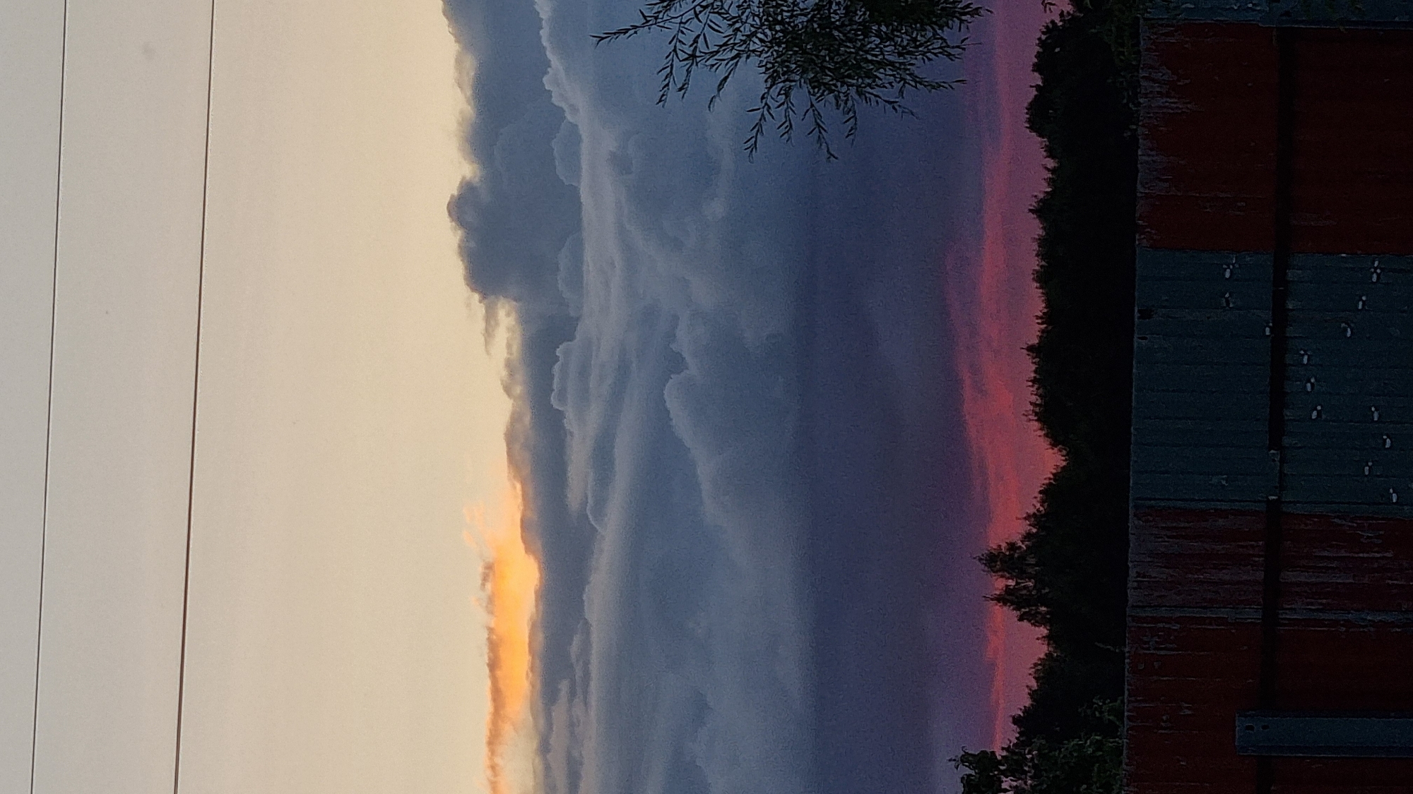 This is a vertical, eye-level, medium shot of a sunset over a rural area, possibly on the outskirts of Vysokoe, Russia. The sky is divided into distinct horizontal bands of color. The top of the frame shows a pale yellow sky, transitioning to a bright orange band of clouds near the horizon. Below this, thick, dark gray clouds dominate the middle of the frame, with lighter gray formations visible within them. The bottom portion of the sky features a deep purple hue. A dark silhouette of trees forms a dense line across the horizon. In the foreground, a weathered fence made of corrugated metal panels, some red and some gray, runs horizontally across the bottom of the image. A single thin electrical wire is visible across the very top of the sky. No people are present in the frame. The overall impression is of a dramatic and moody sunset scene.