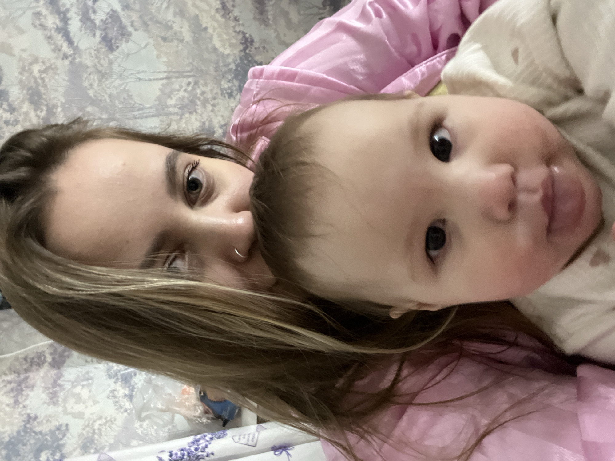 A woman and a baby are lying close together. The woman has long blonde hair and is wearing a pink top. The baby, dressed in white, is looking at the camera with its tongue slightly sticking out. The background appears to be a bed with a patterned bedspread. The location is specified as Novopokrovskaya, Russia, but there is no visible evidence in the image to confirm this location.