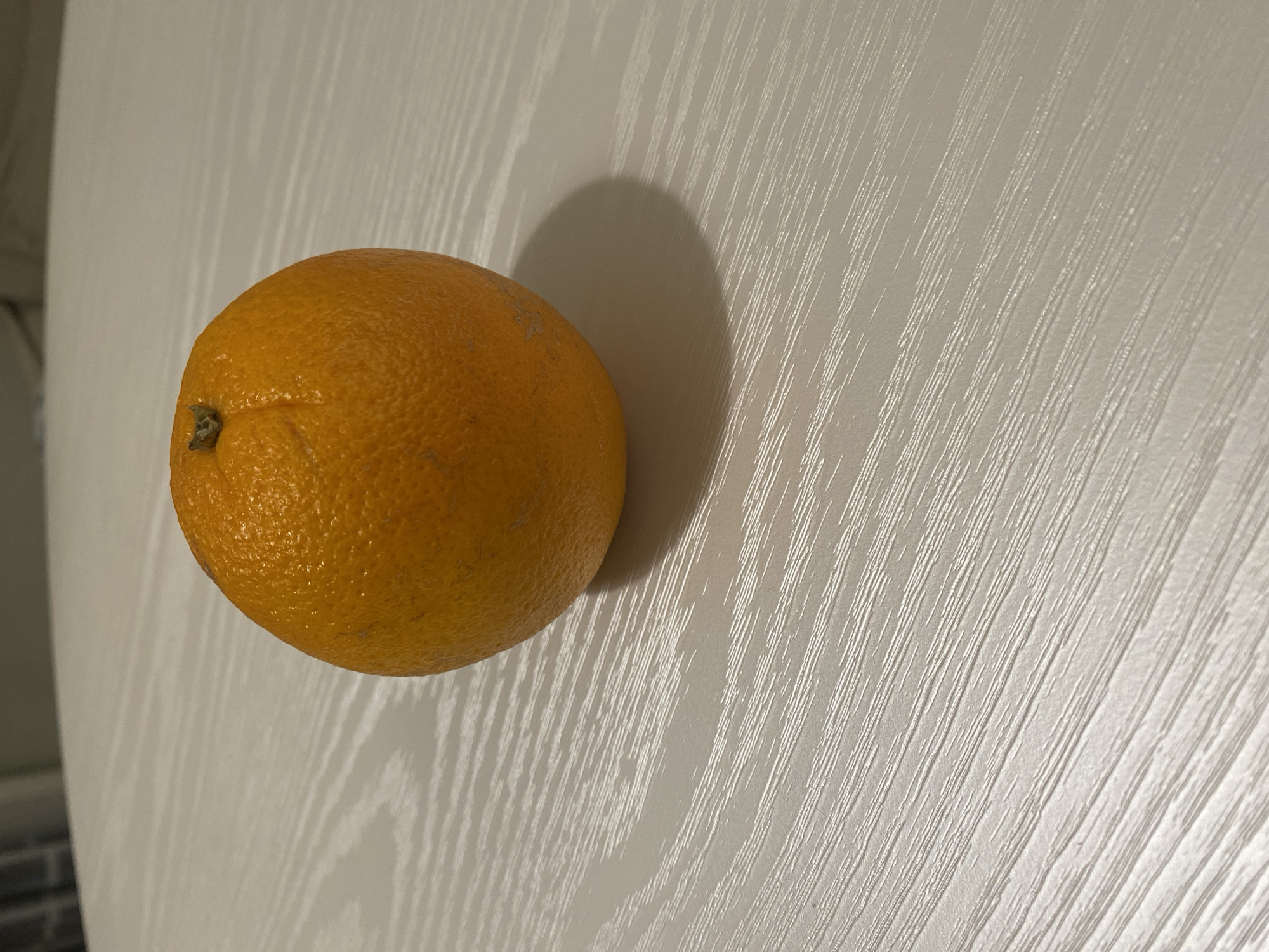 A single, whole, unpeeled orange is positioned on a flat, light-colored surface. The orange has textured, bright orange skin with visible pores and a small, dry, green stem remnant at its top indentation. A distinct shadow is cast by the orange onto the surface, extending towards the lower right, indicating a primary light source from the upper left. The surface itself displays a pronounced, linear wood grain pattern consisting of textured, light-colored striations against a slightly darker, grayish-white background. The background behind the table is out of focus, revealing a blurred, light-toned environment. The scene is located in the city of Unknown, Unknown.