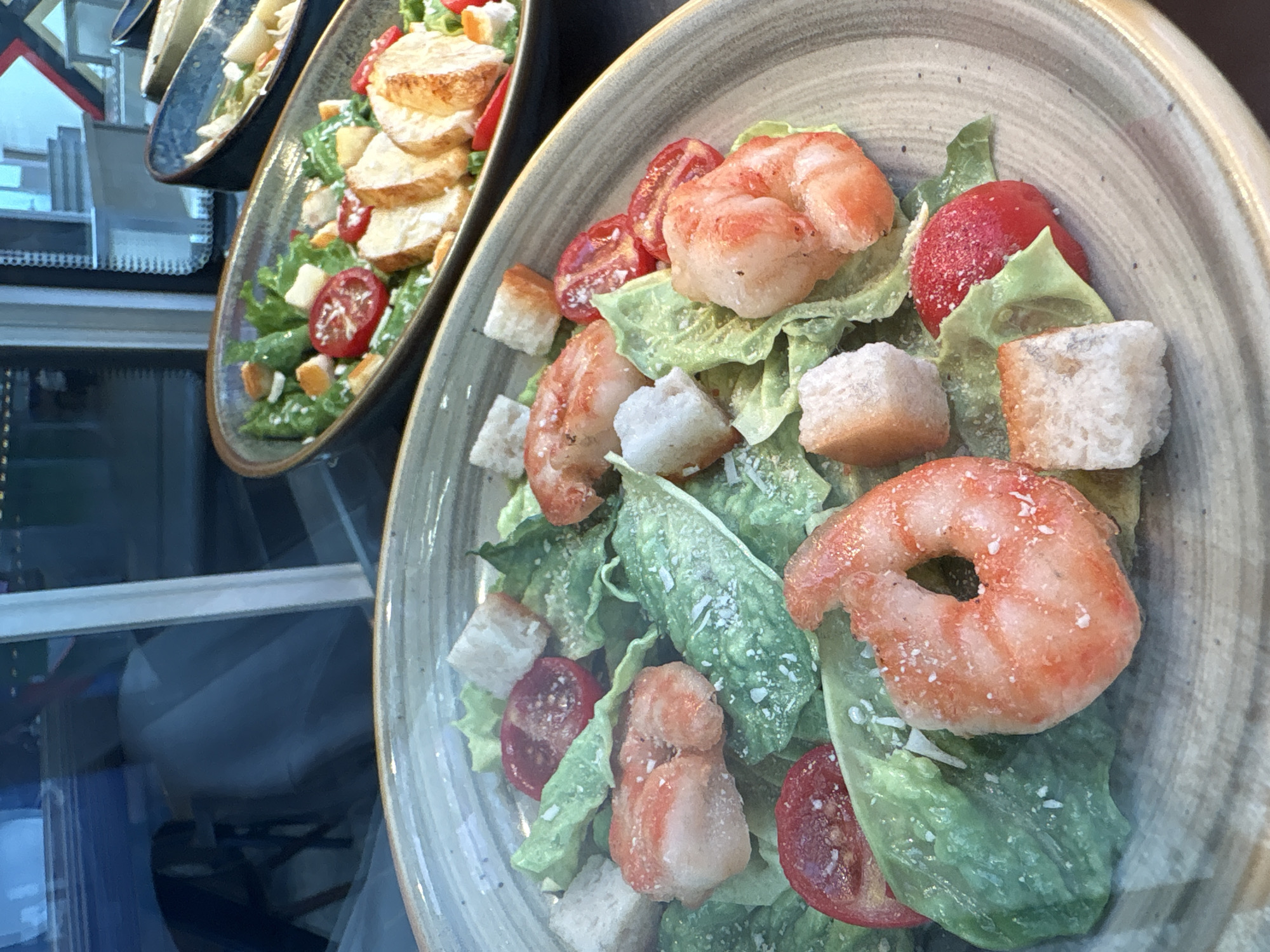 The image shows two plates of shrimp Caesar salad. The salads contain shrimp, romaine lettuce, croutons, and halved tomatoes. The plates are grey with a darker rim, and they appear to be displayed in a restaurant window. The location is Unknown, Unknown.