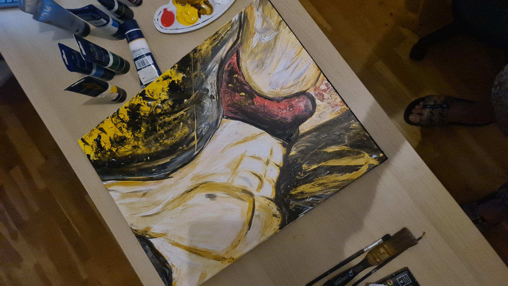 The scene is indoors. A painting of a nude figure lies on a light-colored surface. The painting is executed with yellow, black, red, and white paints. To the left of the painting, there are several tubes of paint and a paint palette. To the right of the painting, there are paintbrushes. In the background, there is a person wearing sandals and sitting on a wooden floor. There is nothing to indicate this image was taken in Budva, Montenegro.